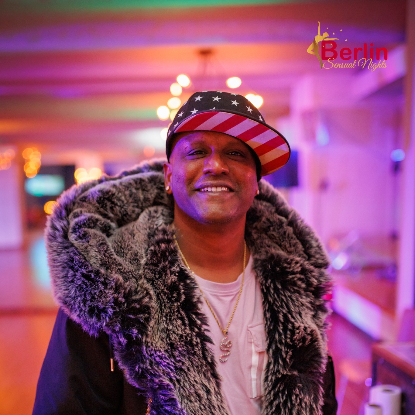 Smiling man wearing a fur coat and a patriotic cap in a colorful indoor setting.