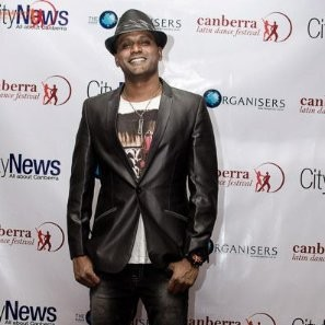 Man in black blazer, hat, and casual clothing smiling at event with backdrop featuring logos and text.