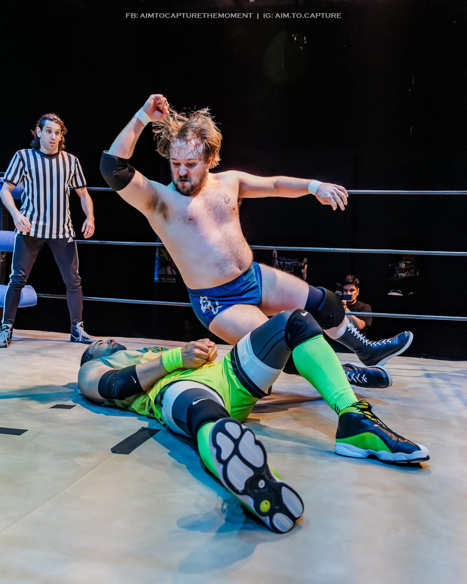 Wrestler in blue shorts and black hair strikes a pose over an opponent in green and black gear on the ring floor.