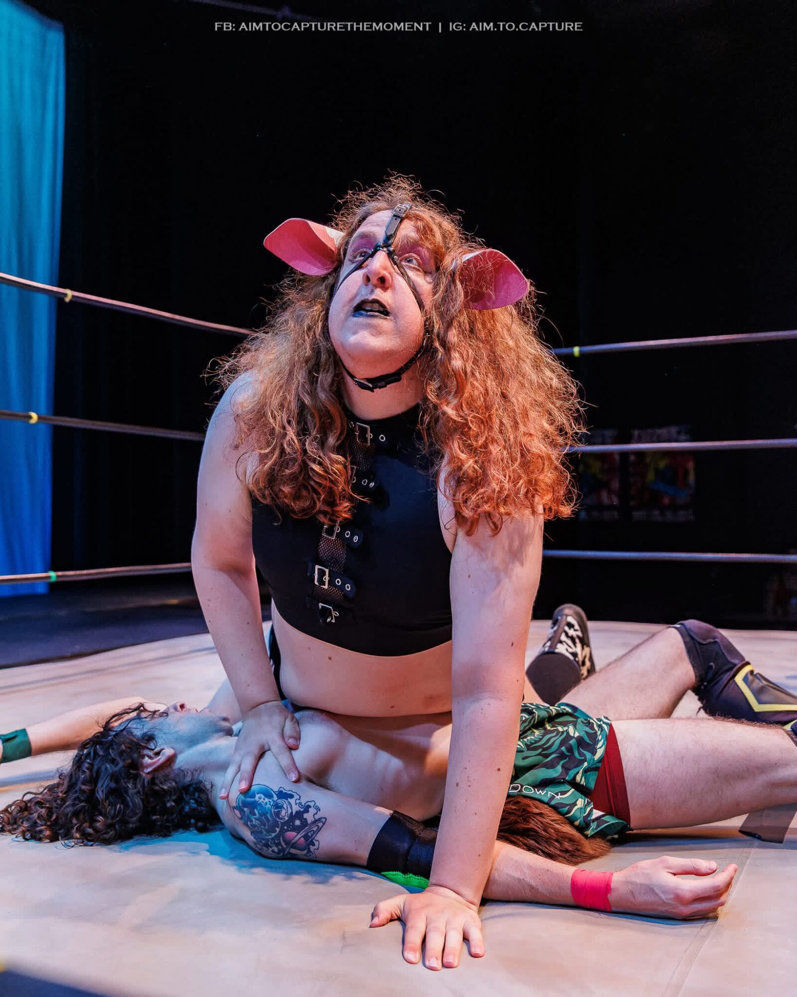 Wrestling match with two women, one on top, in a ring with ropes, one lying on the mat, with a blue curtain background.