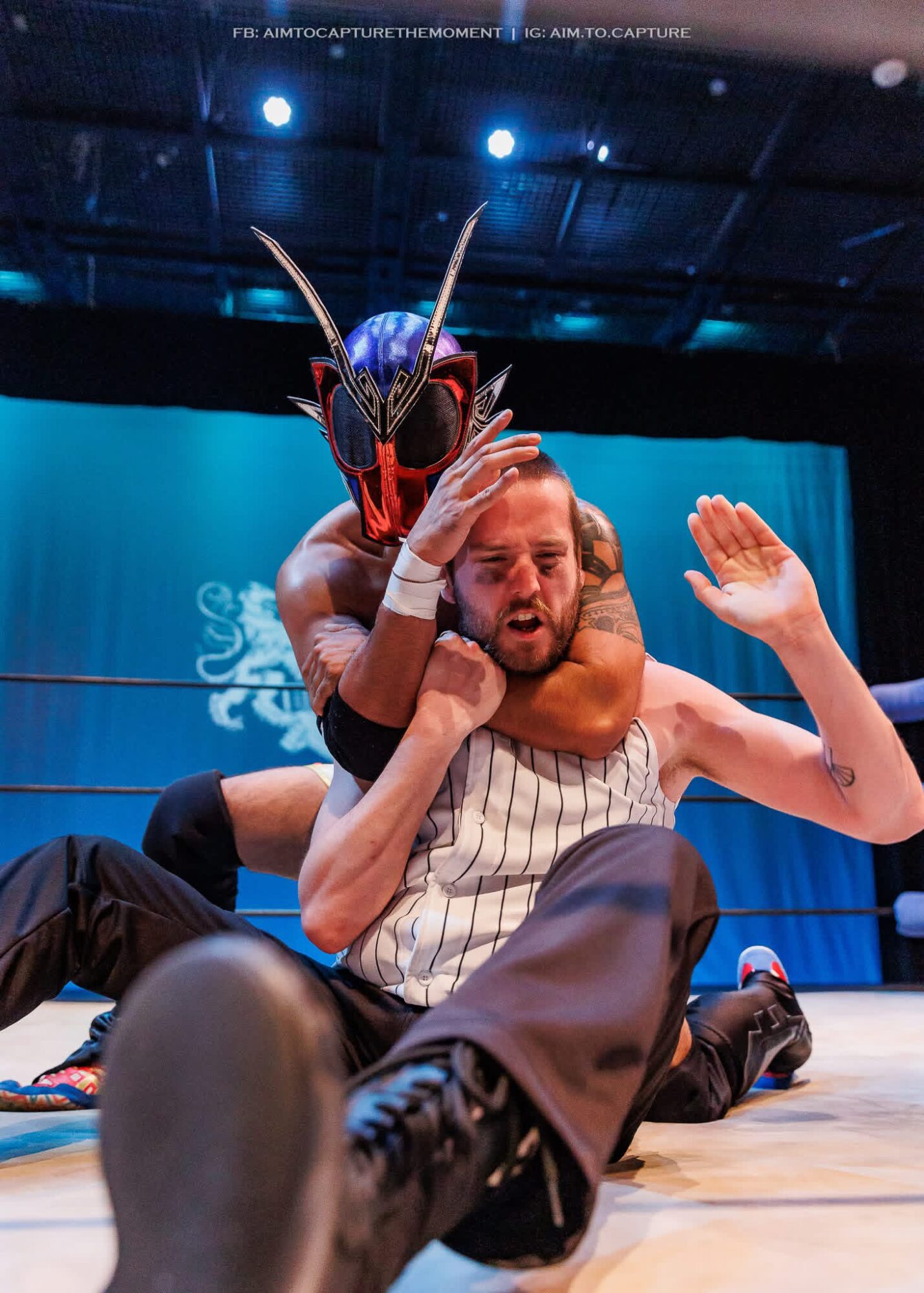 Wrestler wearing a mask with antennae holds a man in a striped shirt in a wrestling ring, with a blue background.
