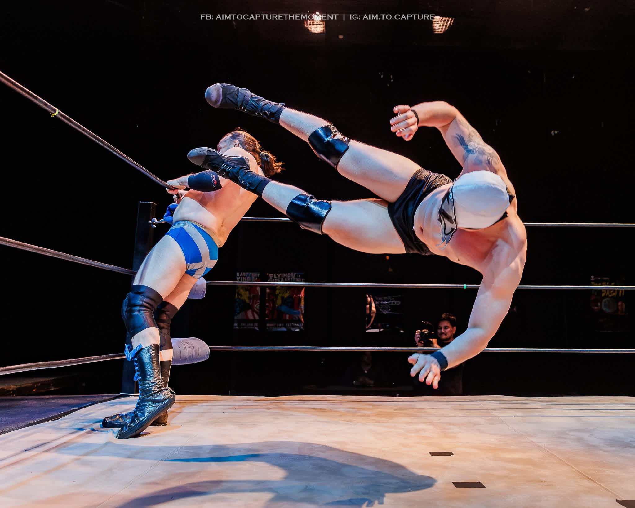 Two wrestlers in a ring, one delivering a kick to the other's head, with ropes visible in the background.