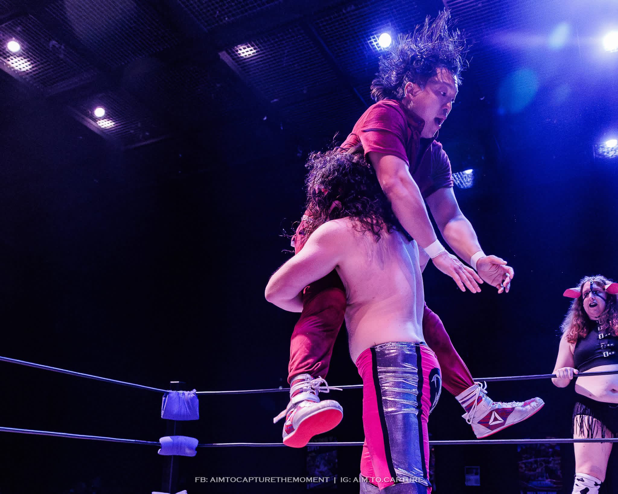 Two wrestlers in a ring, one lifting the other, with a referee and a woman in the background, under bright lights.
