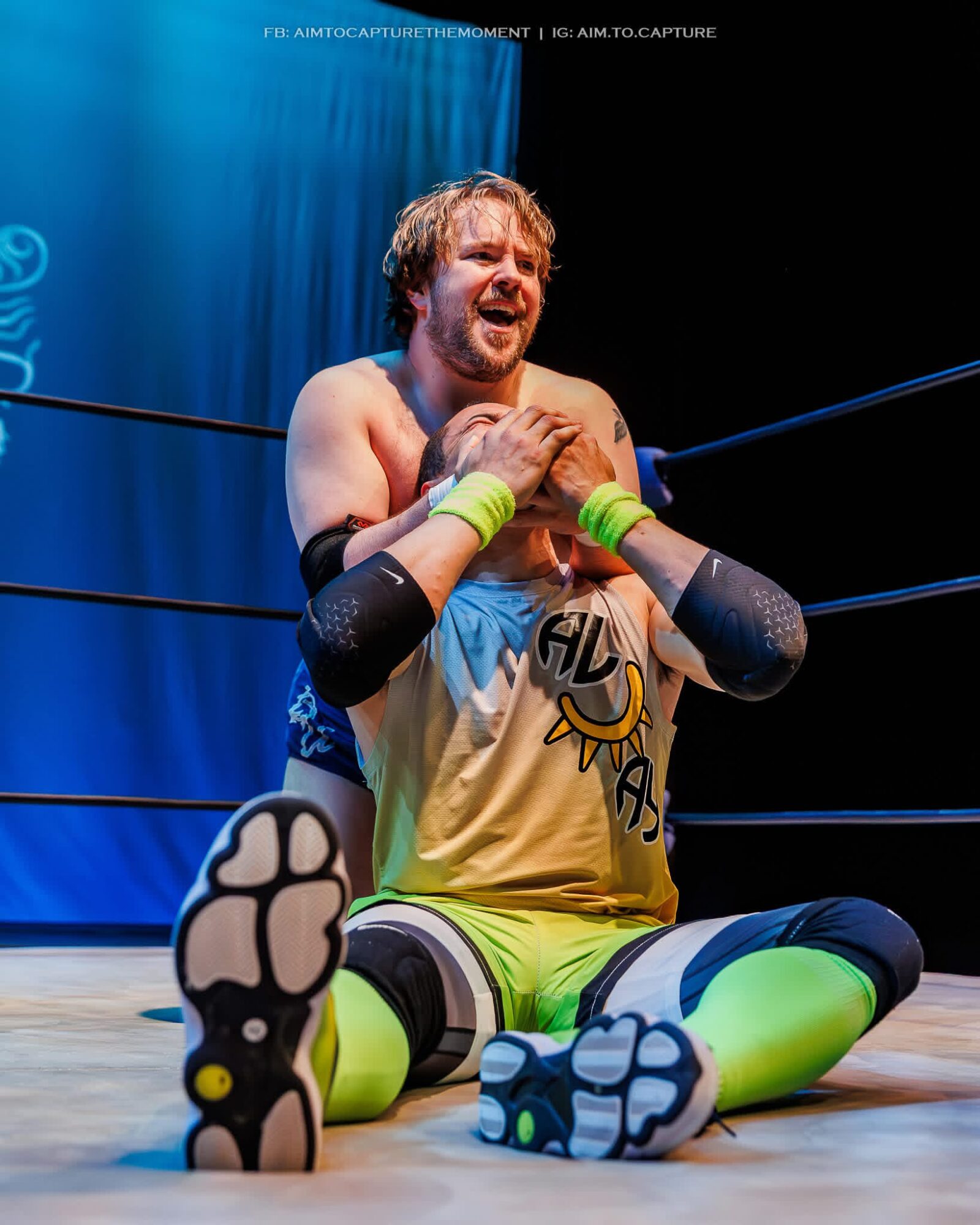 Two wrestlers in ring, one sitting and crying, the other embracing him, with blue curtain background.