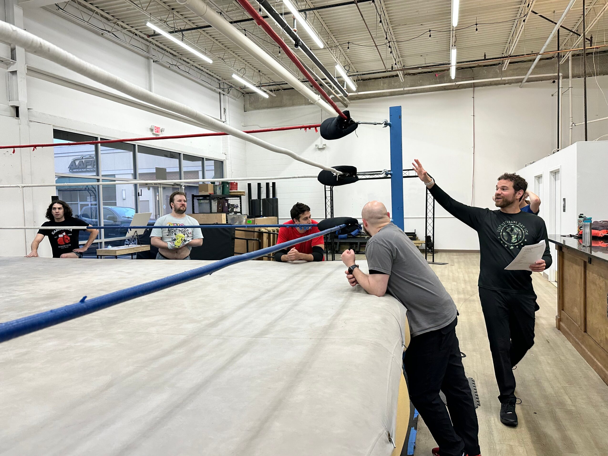 People in a boxing gym with a ring, some sitting and one standing and talking, in a spacious room with large windows.
