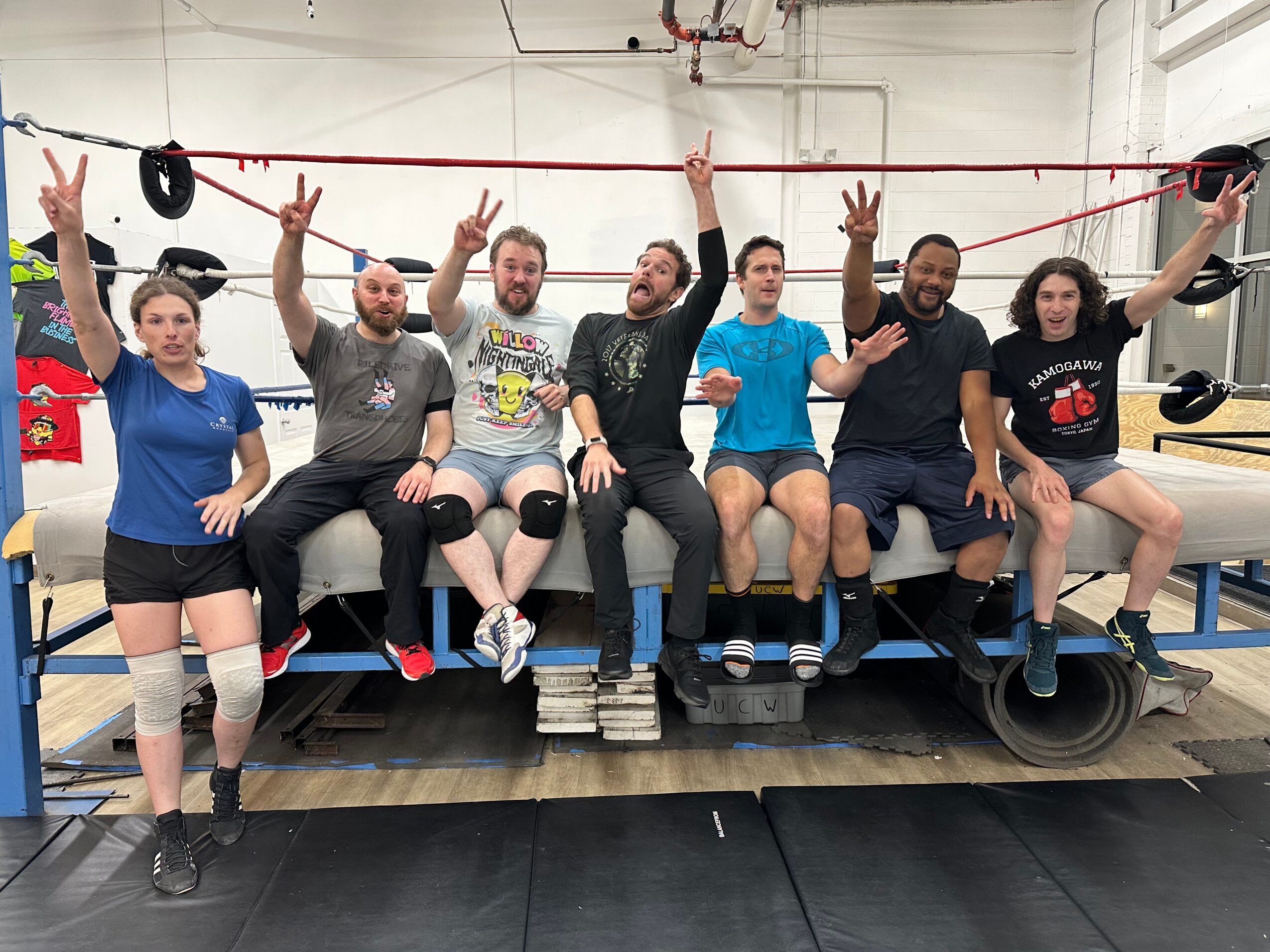 Group of seven people sitting on a bench in a gym, raising their hands, with gym equipment in background.