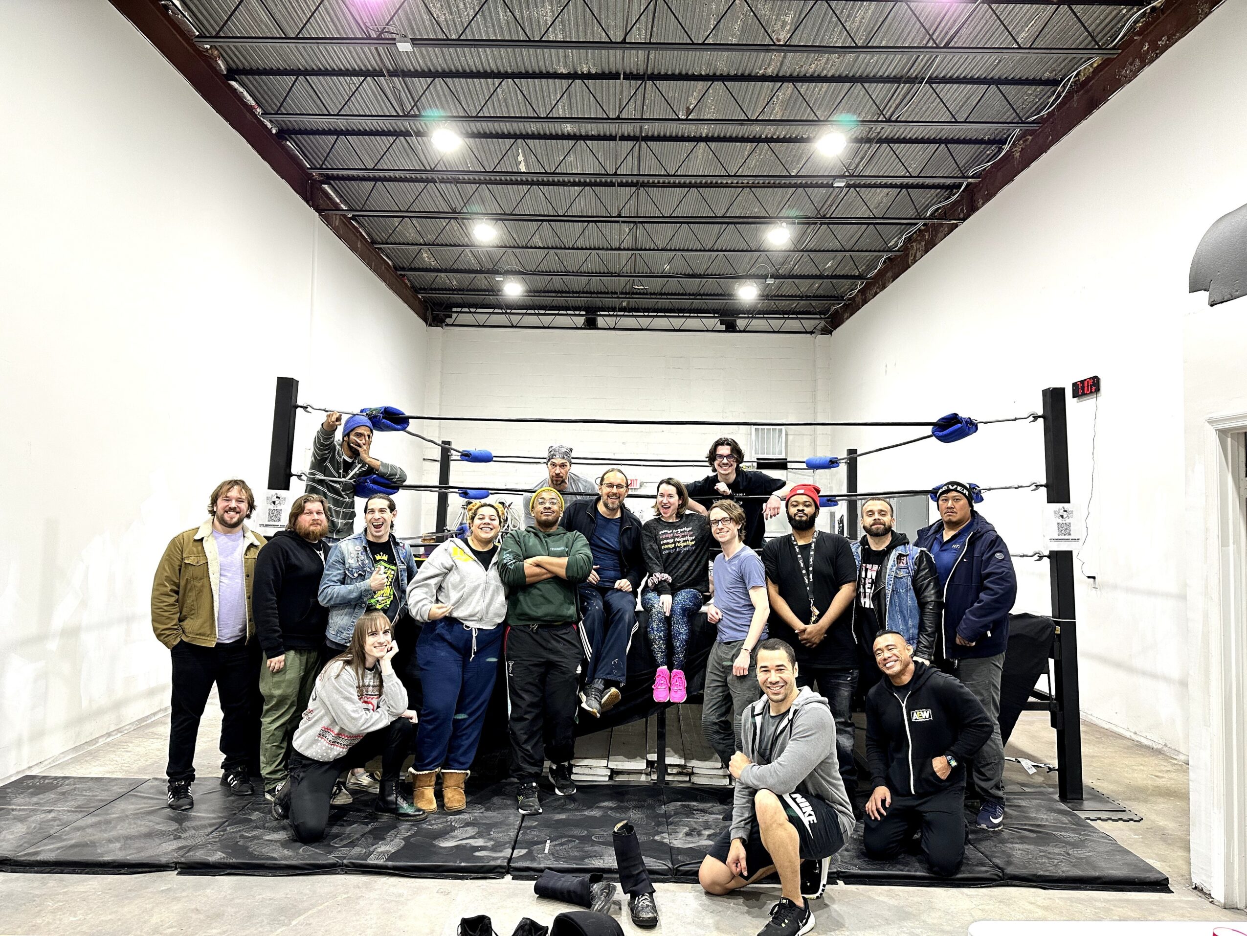 Group of people in a boxing ring inside a room with white walls and a high ceiling, some smiling and posing.