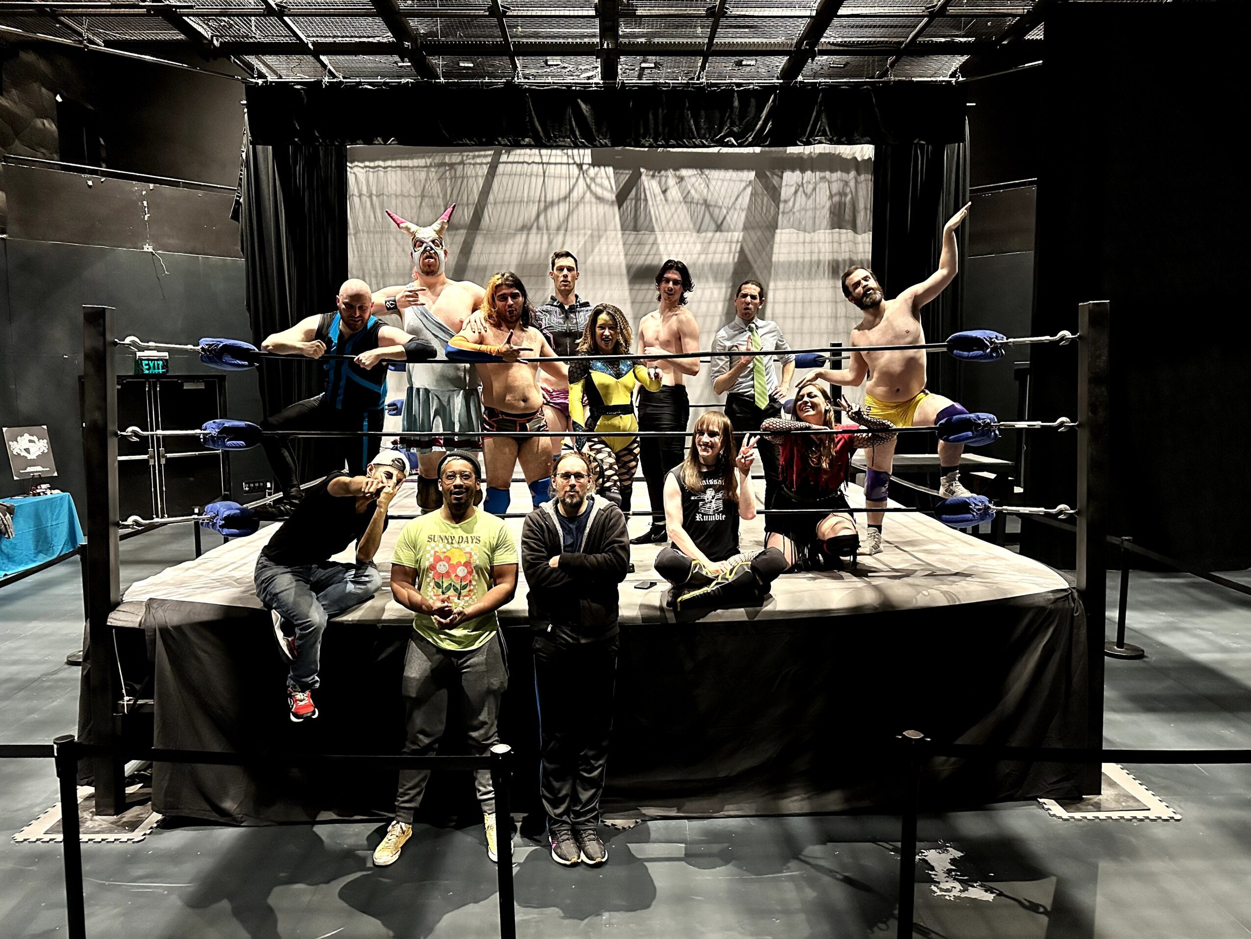 Group of people in a wrestling ring, some with costumes, posing and smiling, in a dark indoor setting.