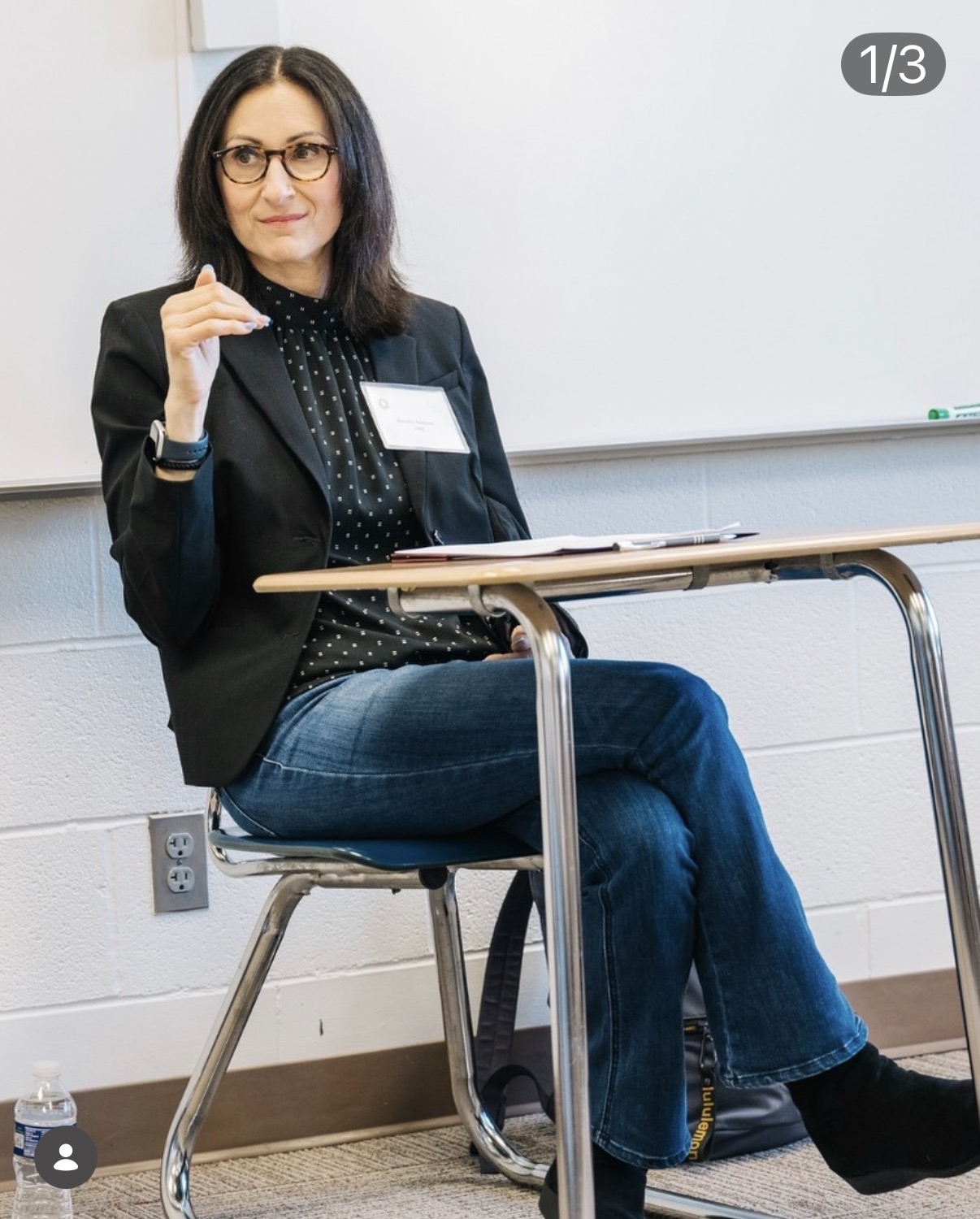 Woman sitting on a chair in a classroom, wearing glasses, a black blazer, and jeans, with a whiteboard behind her.