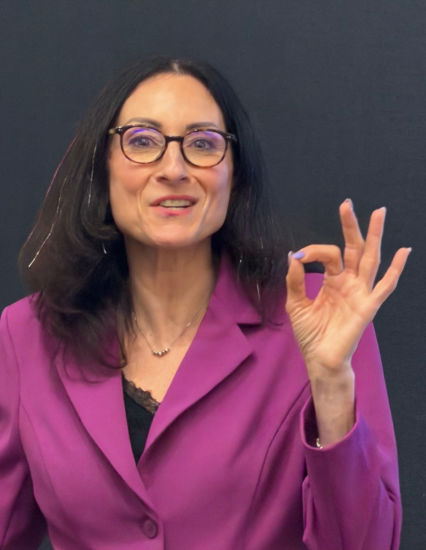 Woman with glasses and dark hair wearing a purple blazer, making an OK gesture with her right hand.