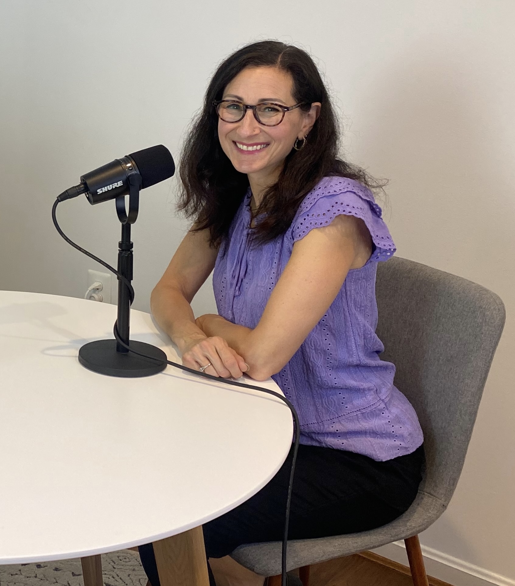 Woman with glasses smiling, sitting at a table with a microphone, wearing a purple top, in a room with white walls.
