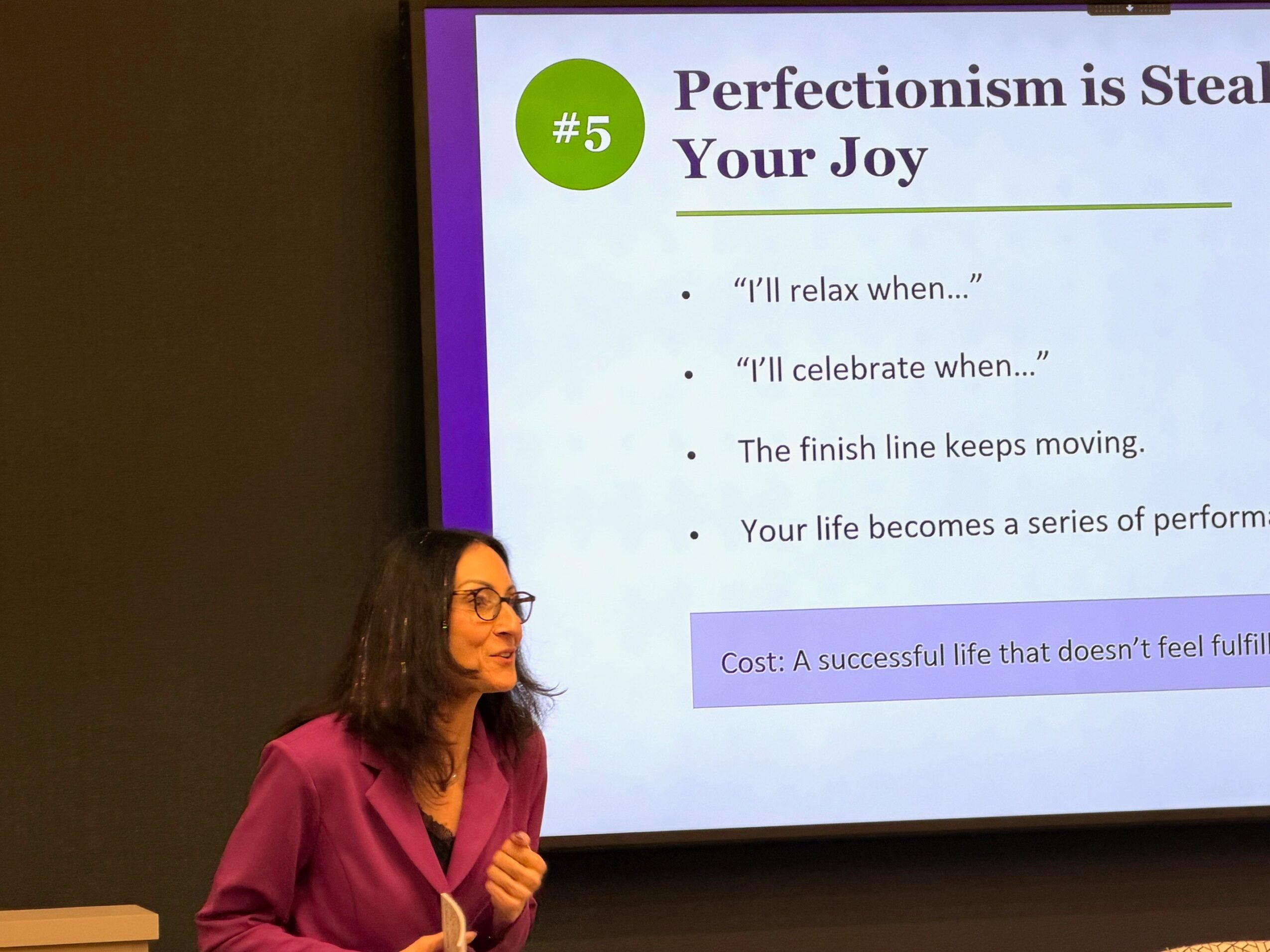 Woman with glasses and curly hair in a purple blouse standing in front of a presentation slide with text about perfectionism and joy.