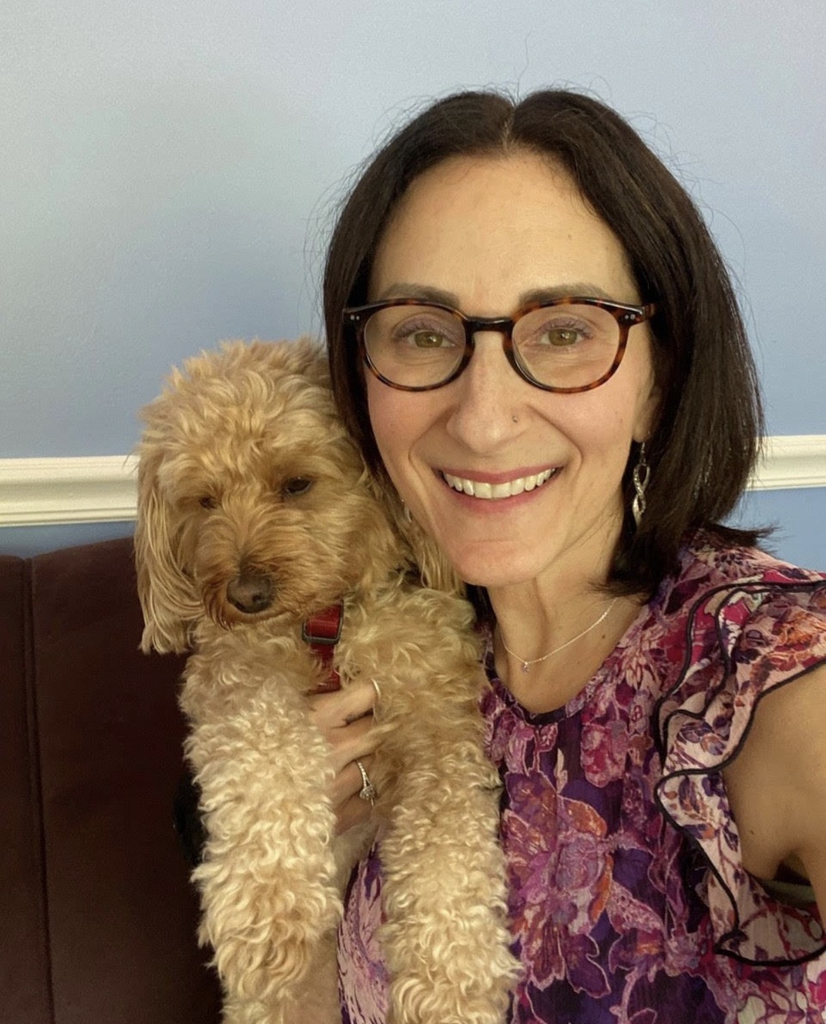 Woman with glasses smiling, holding a curly-haired dog, in front of a blue wall.