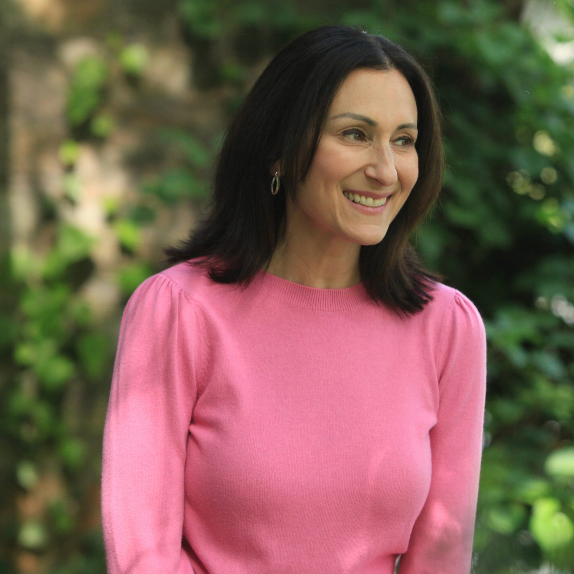 Woman with dark hair smiling outdoors, wearing a pink top, surrounded by green foliage.