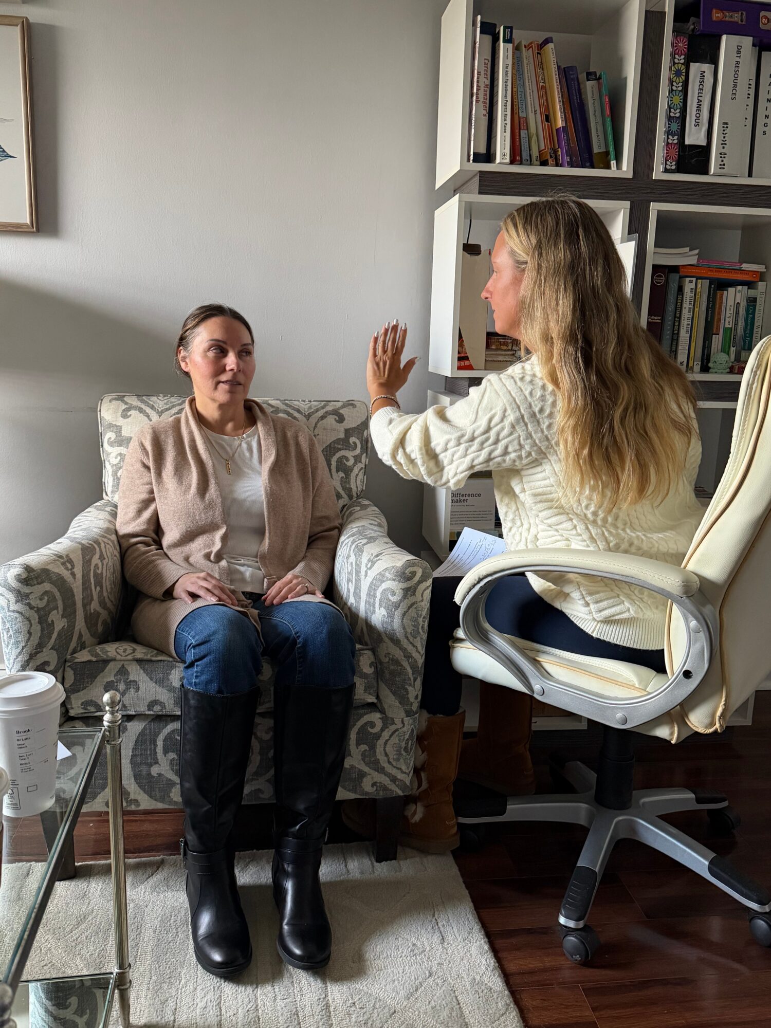 Two women engaged in conversation in a cozy room with bookshelves, one sitting on a sofa and the other in a chair.