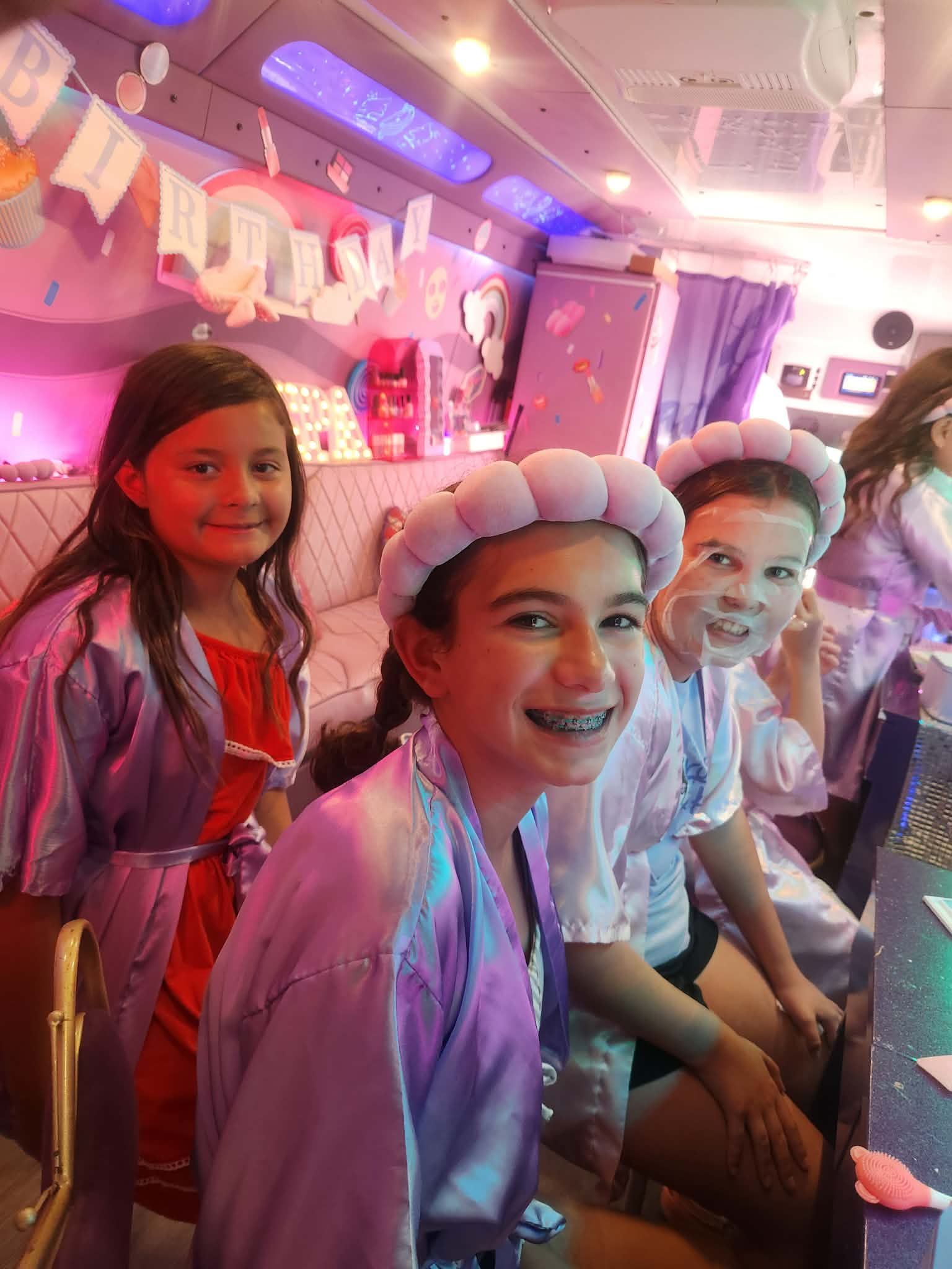 Three children smiling at a party, one wearing a chef's hat, in a decorated room with pink and purple lighting.