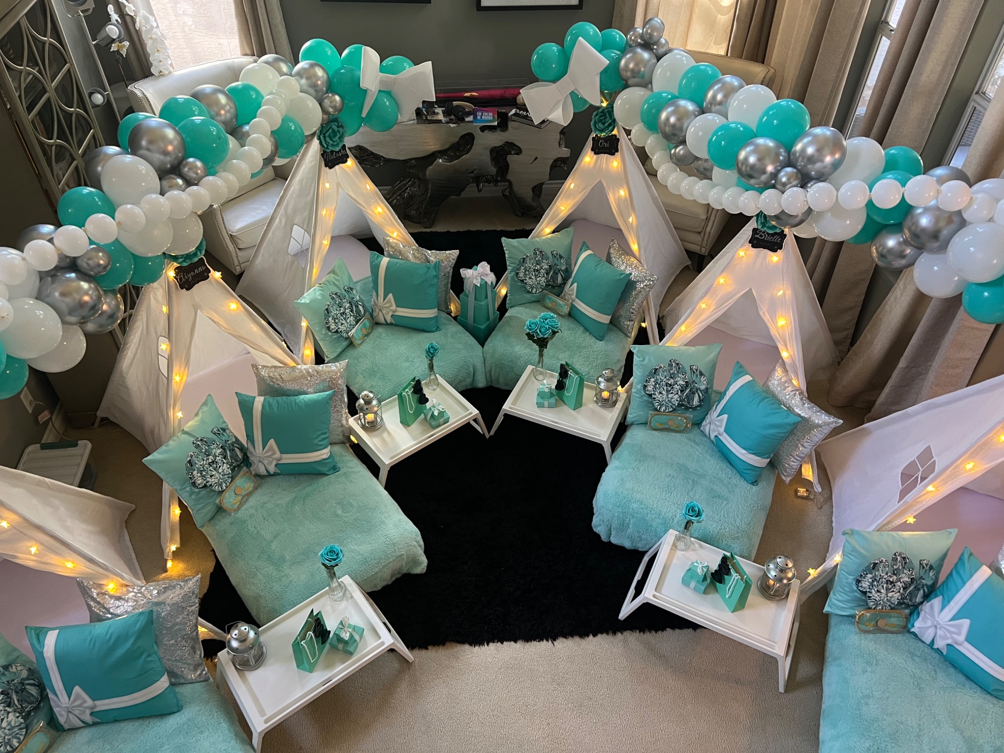 Decorated seating area with teal and white balloons, small tents, and pillows, arranged in a semi-circle around a black rug.