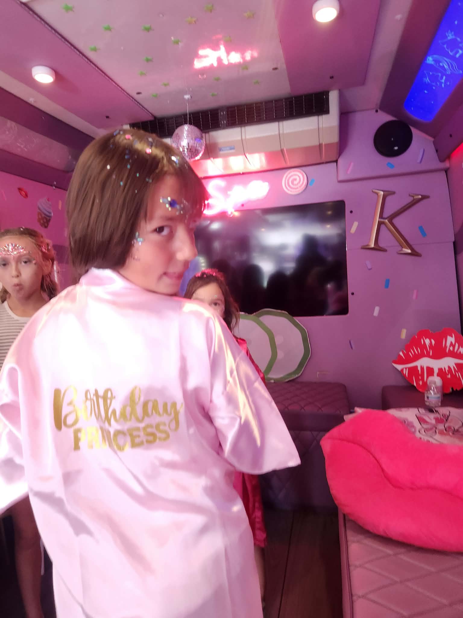 Person with short hair wearing a pink satin robe with gold text, in a decorated room with pink and purple decor, a TV, and a large red plush chair.