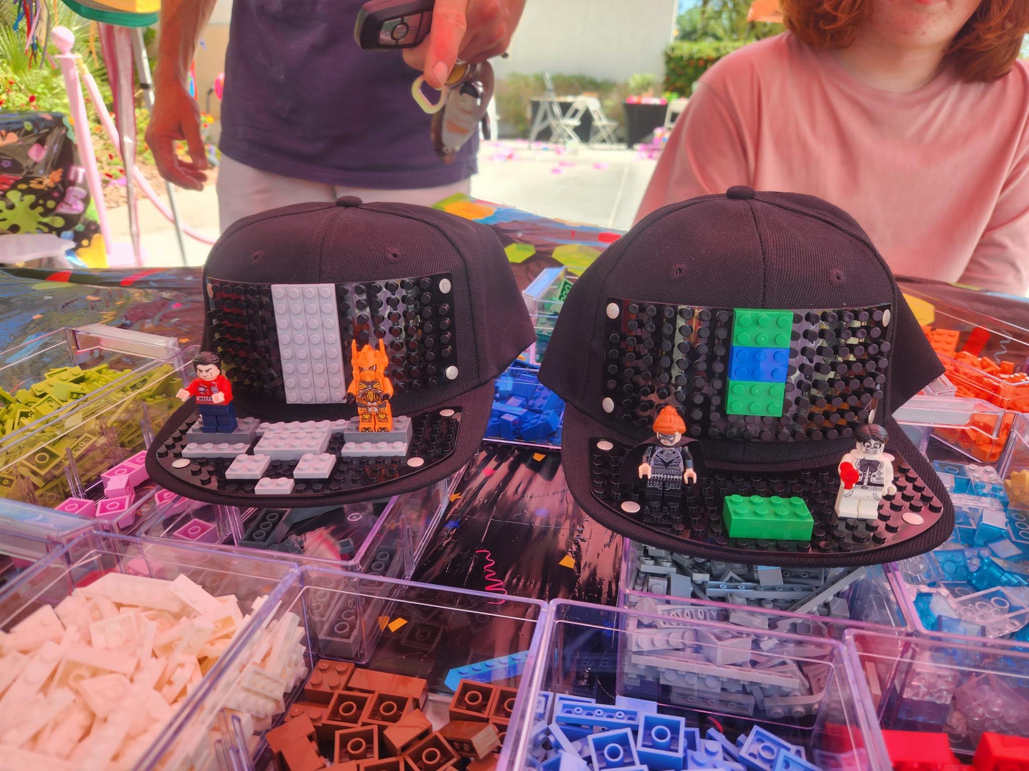 Two open black cases with LEGO minifigures and bricks on a table outdoors, with people in the background.