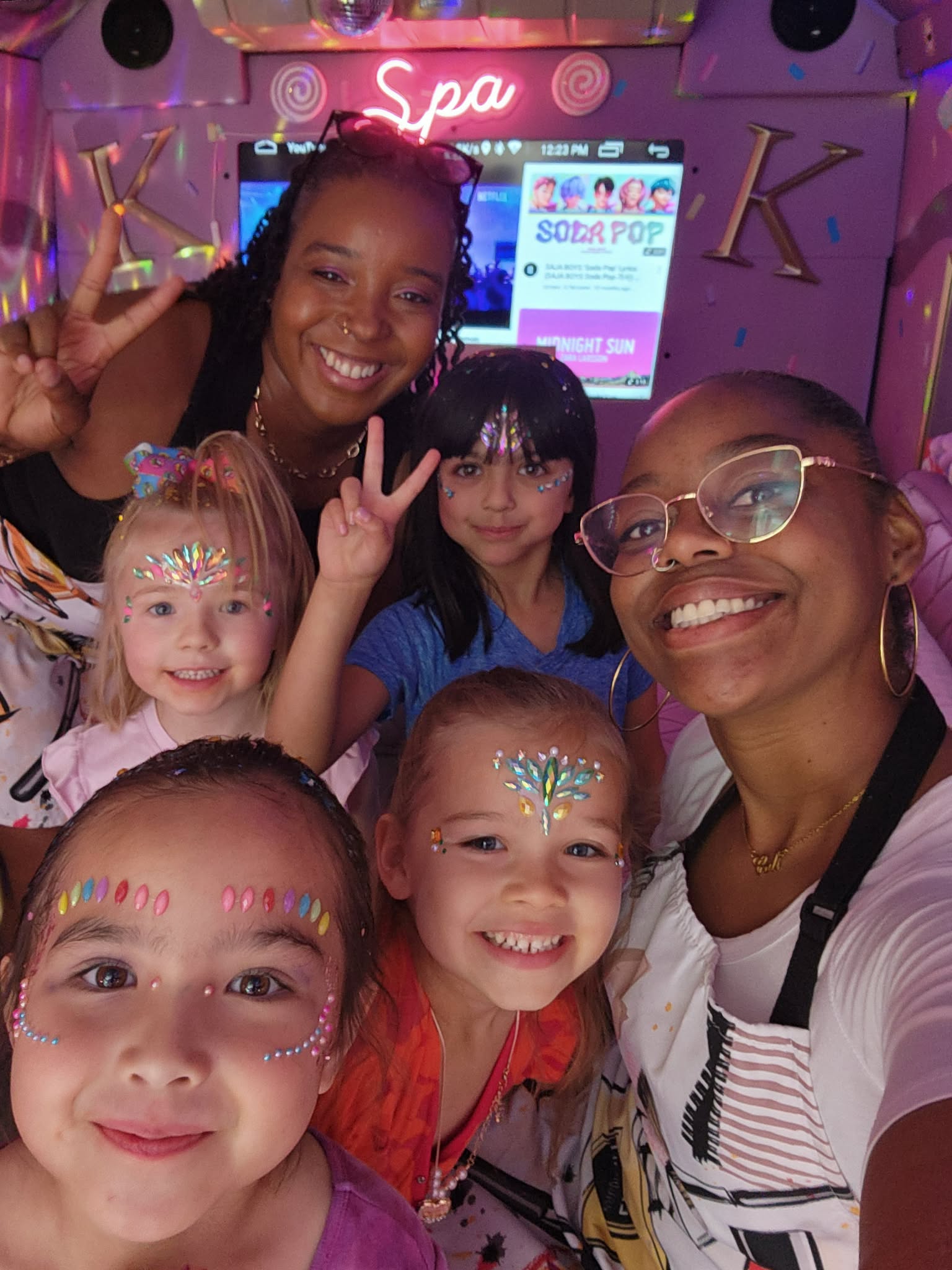 Group of five smiling children and two women with face paint, making peace signs inside a spa with pink decor.