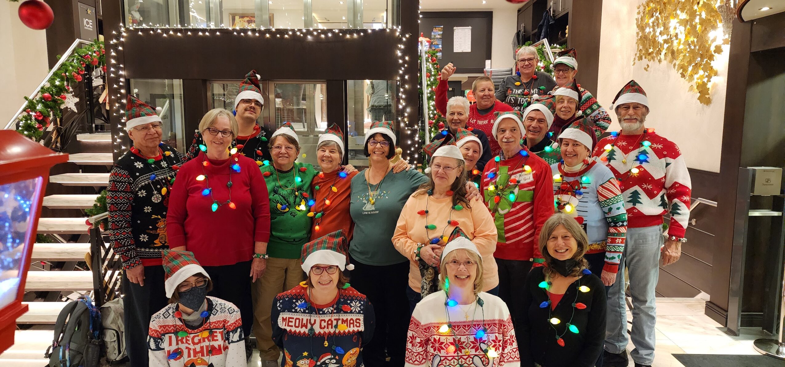 Group of people celebrating Christmas, wearing festive sweaters and hats, indoors with holiday decorations and lights.