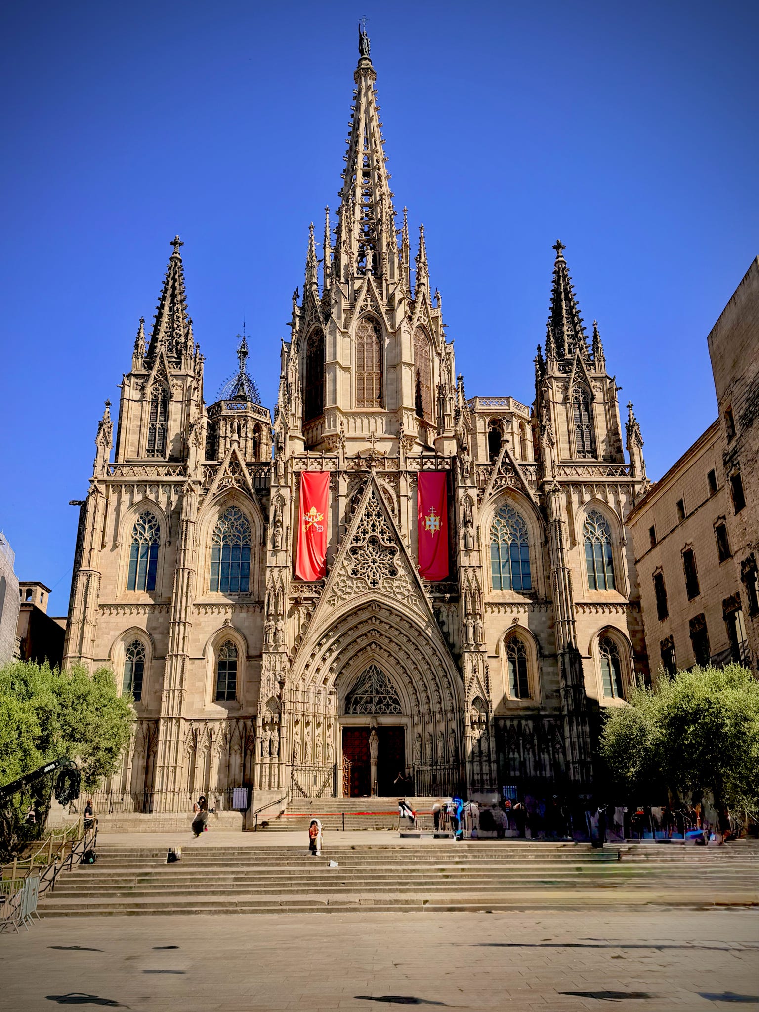 Gothic cathedral with tall spires, ornate facade, and flags hanging near entrance, under a clear blue sky.