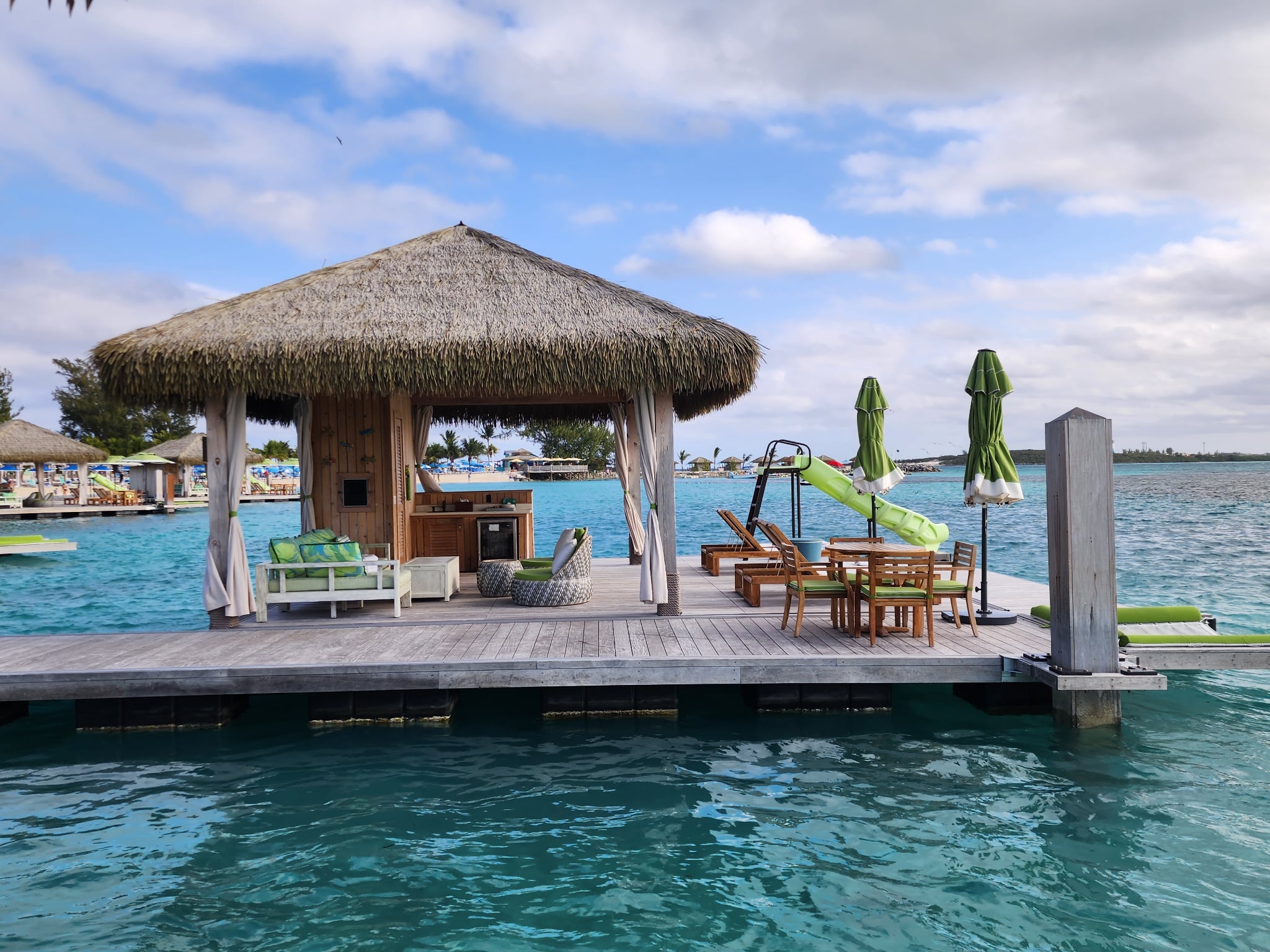 Thatched-roof hut on a dock over water with seating and a slide, under partly cloudy sky.