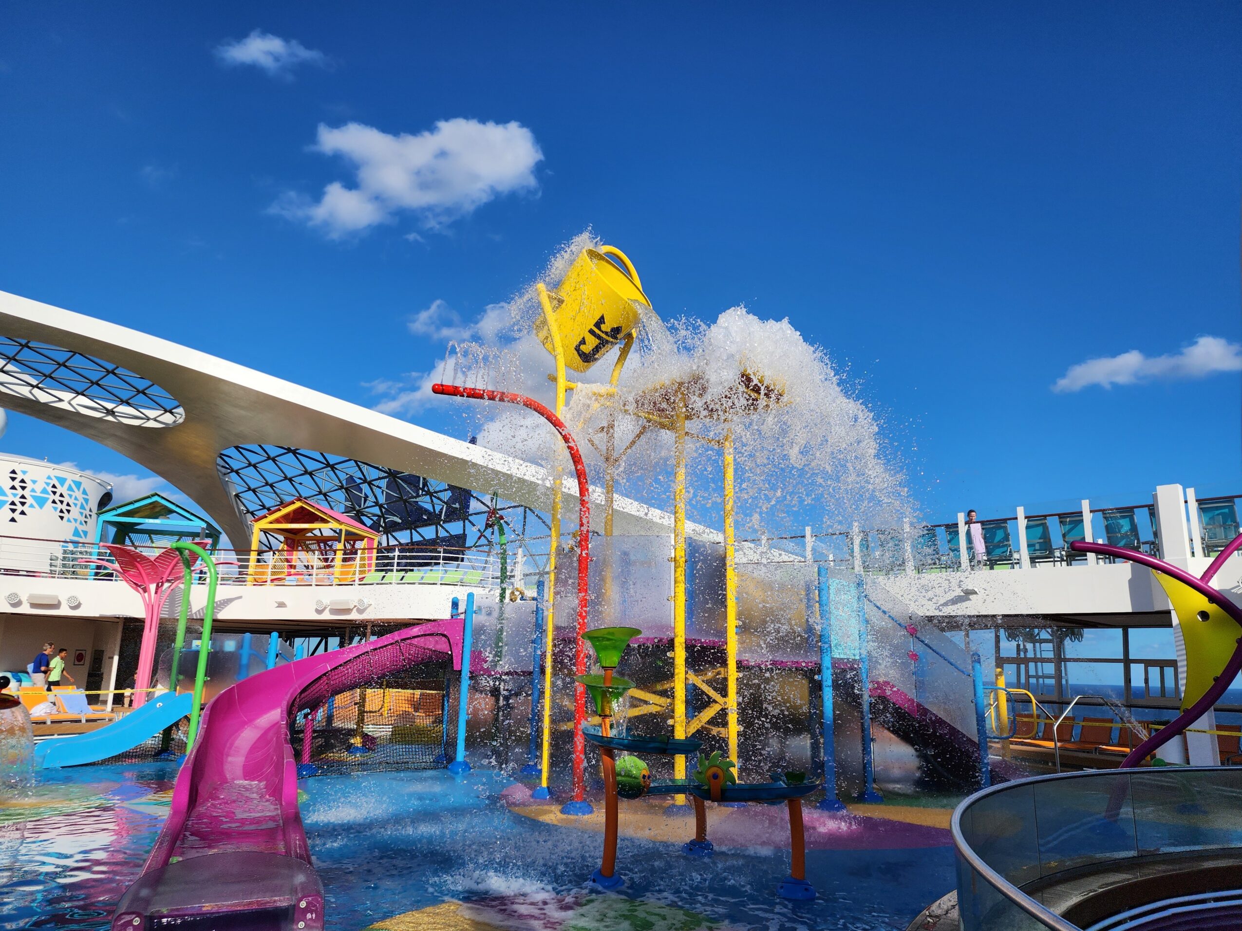 Water park with slides, water sprays, and a yellow bucket pouring water, under a blue sky with clouds.