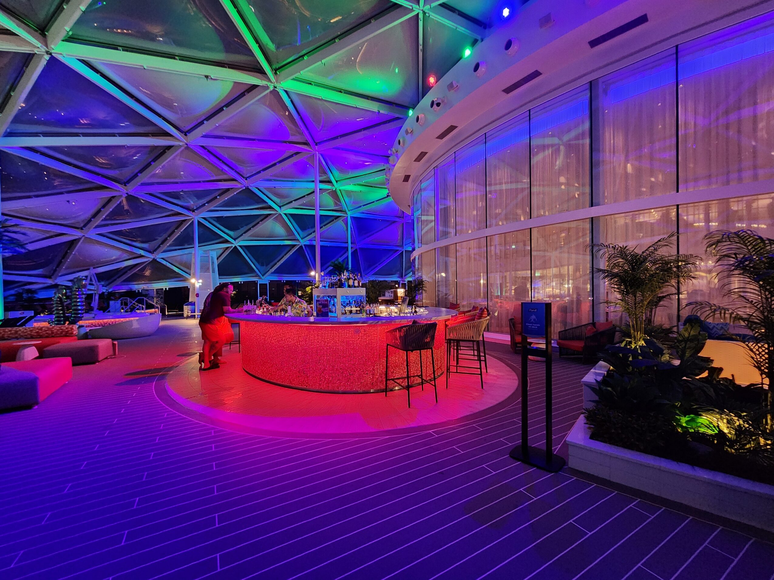 Indoor space with colorful lighting, curved bar, seating area, and large glass windows with plants.