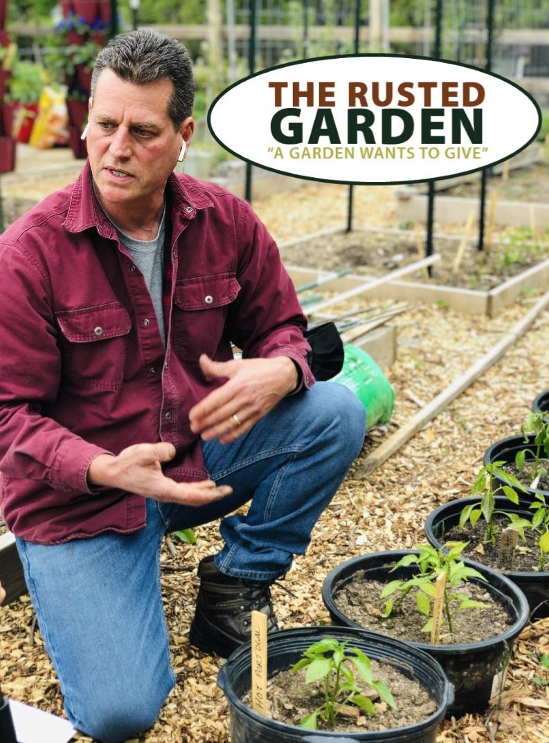 Man kneeling in a garden with potted plants, holding a gardening tool, with a sign reading 'The Rusted Garden' in the background.