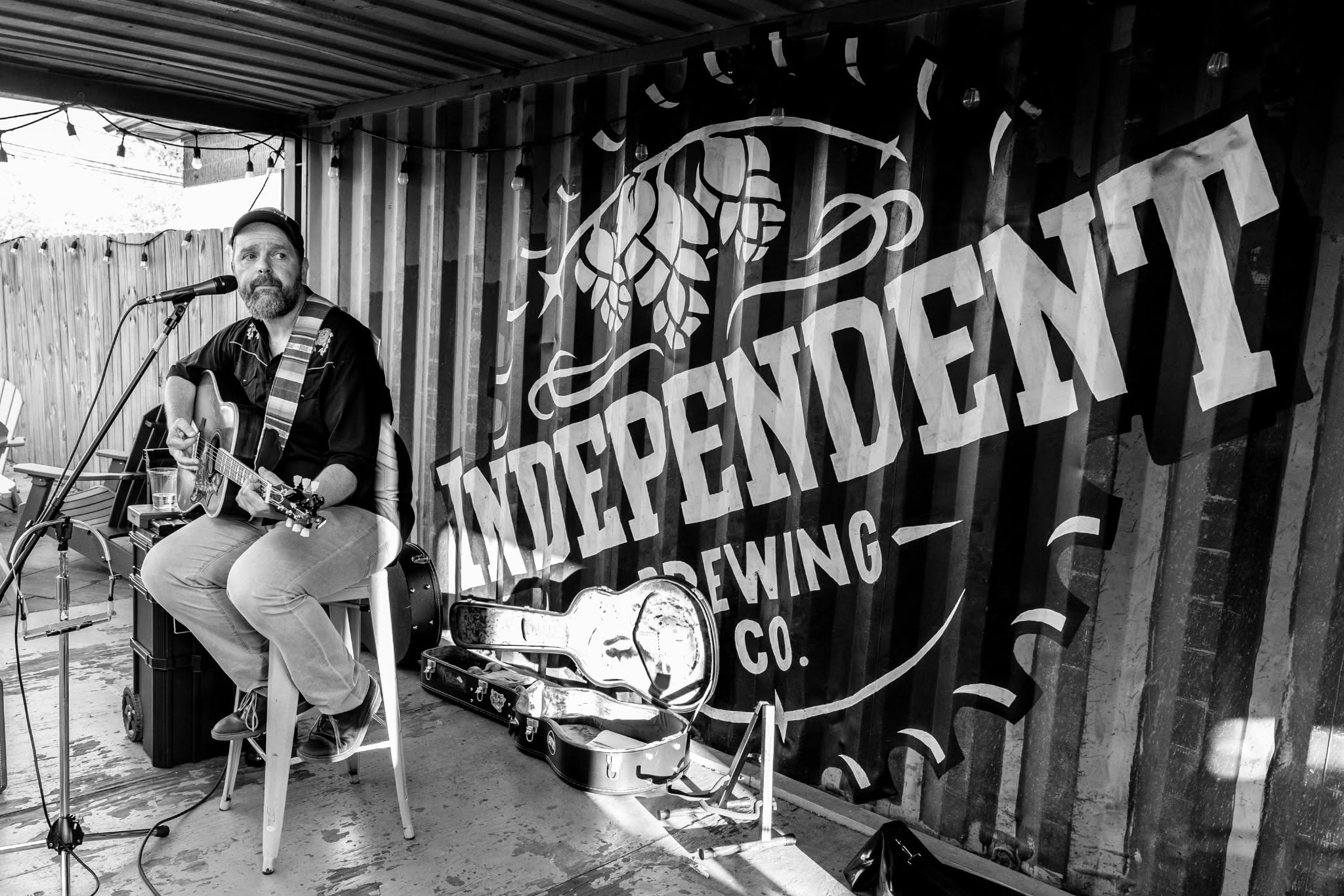 A man plays guitar and sings into a microphone on a small stage with a large 'Independent Brewing Co.' sign behind him.