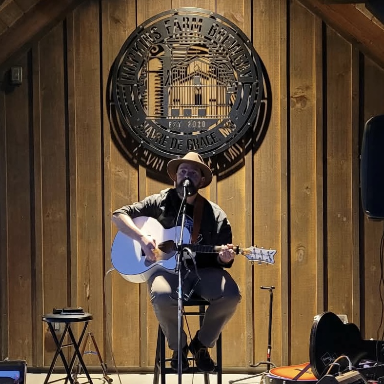 Person playing guitar and singing on stage with wooden background and circular sign above, holding a hat, with a microphone. 20 words.