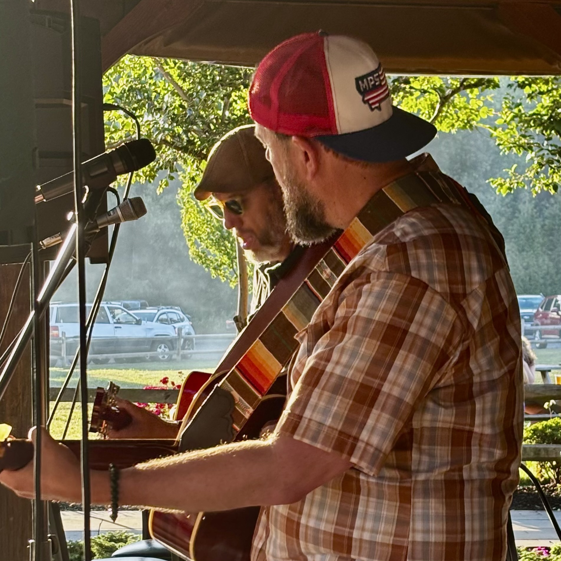 Two men playing guitars on stage, one wearing a red and white cap, outdoors with trees and parked cars in background.