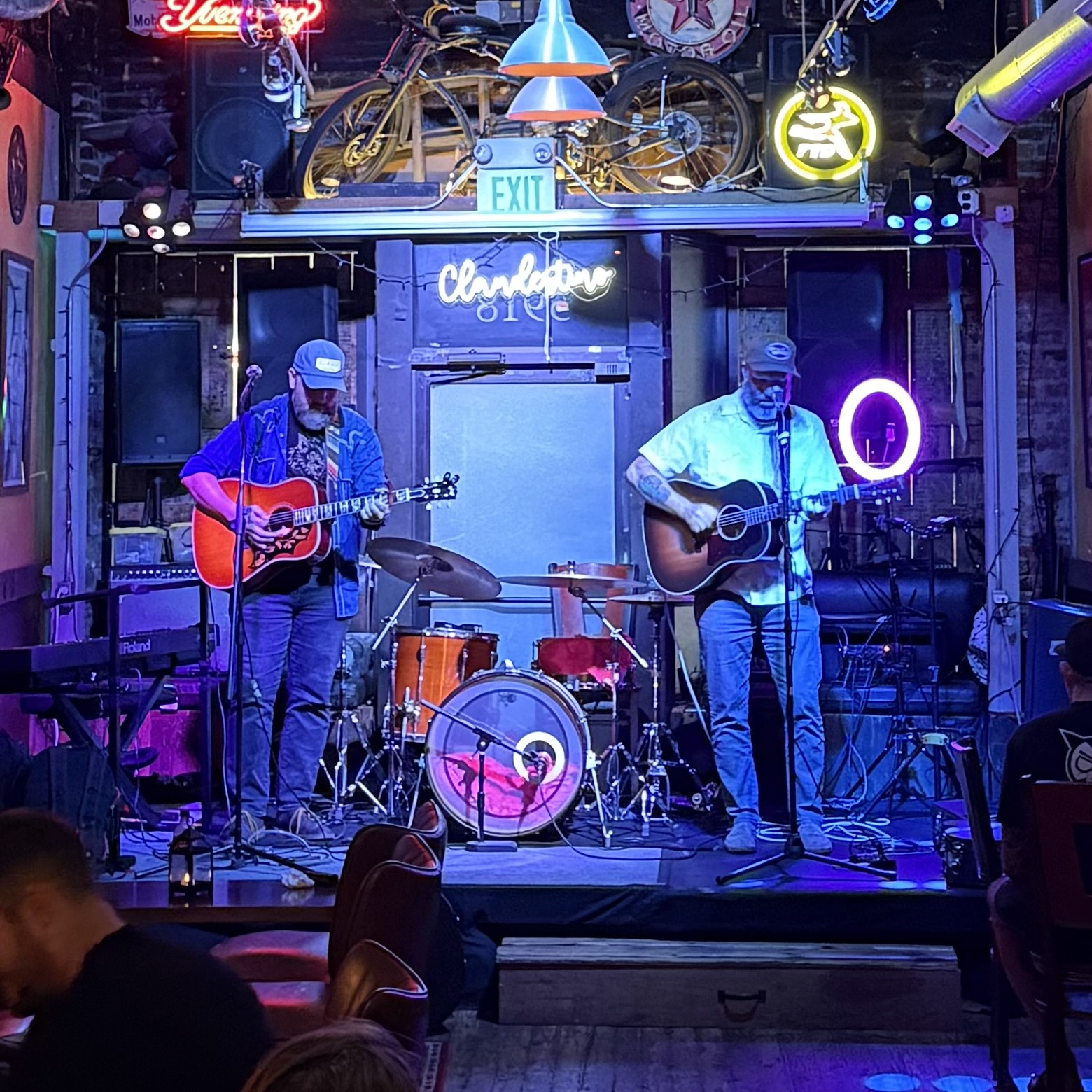 Two musicians playing guitars on a stage with colorful neon signs and a drum set in a dimly lit venue.