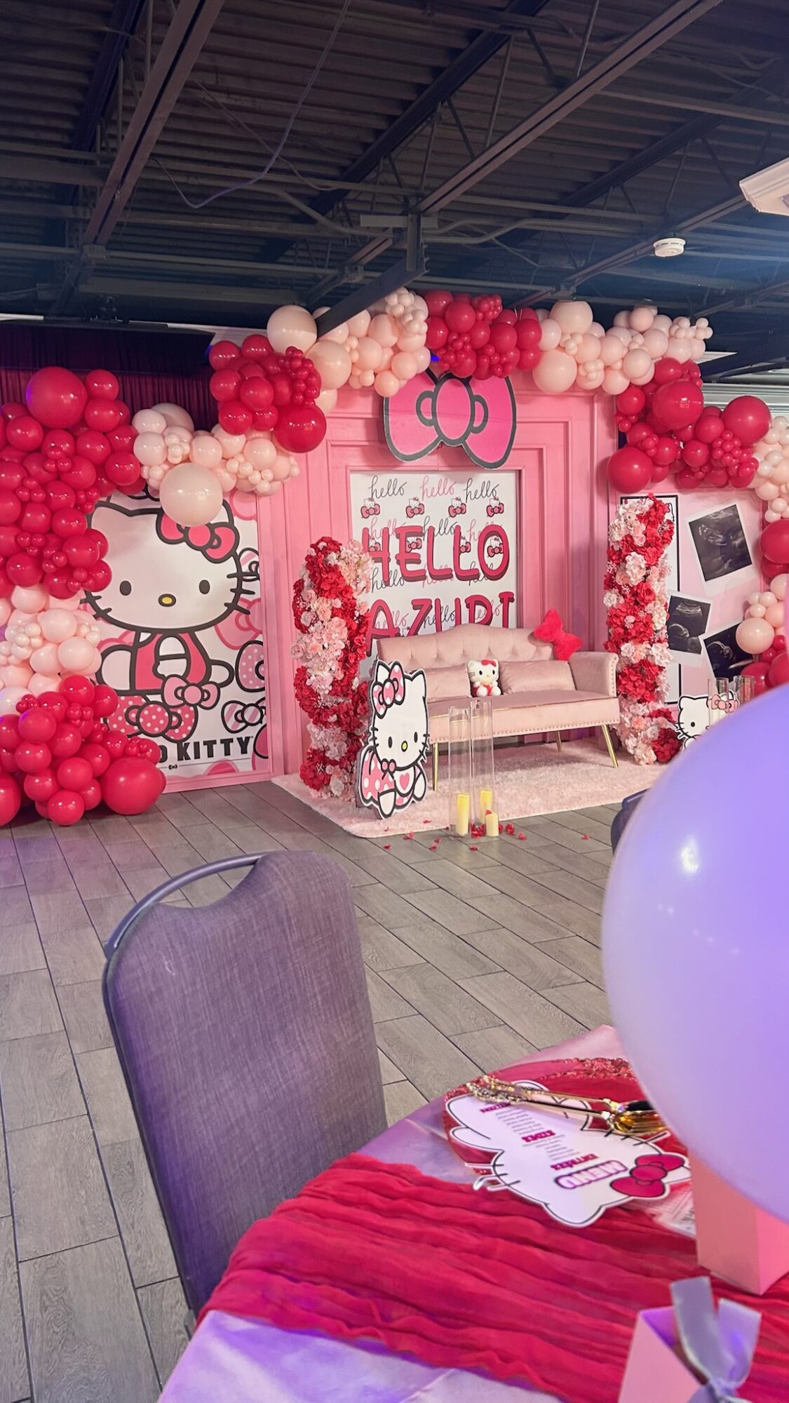 Party decoration with Hello Kitty theme, pink and red balloons, and a backdrop with Hello Kitty and text, in an indoor setting.