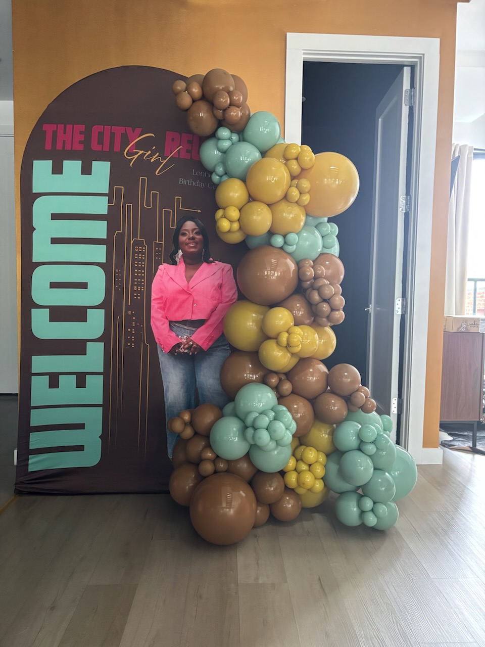 Person standing next to a colorful balloon arrangement and a welcome sign, indoors with a door and window nearby.