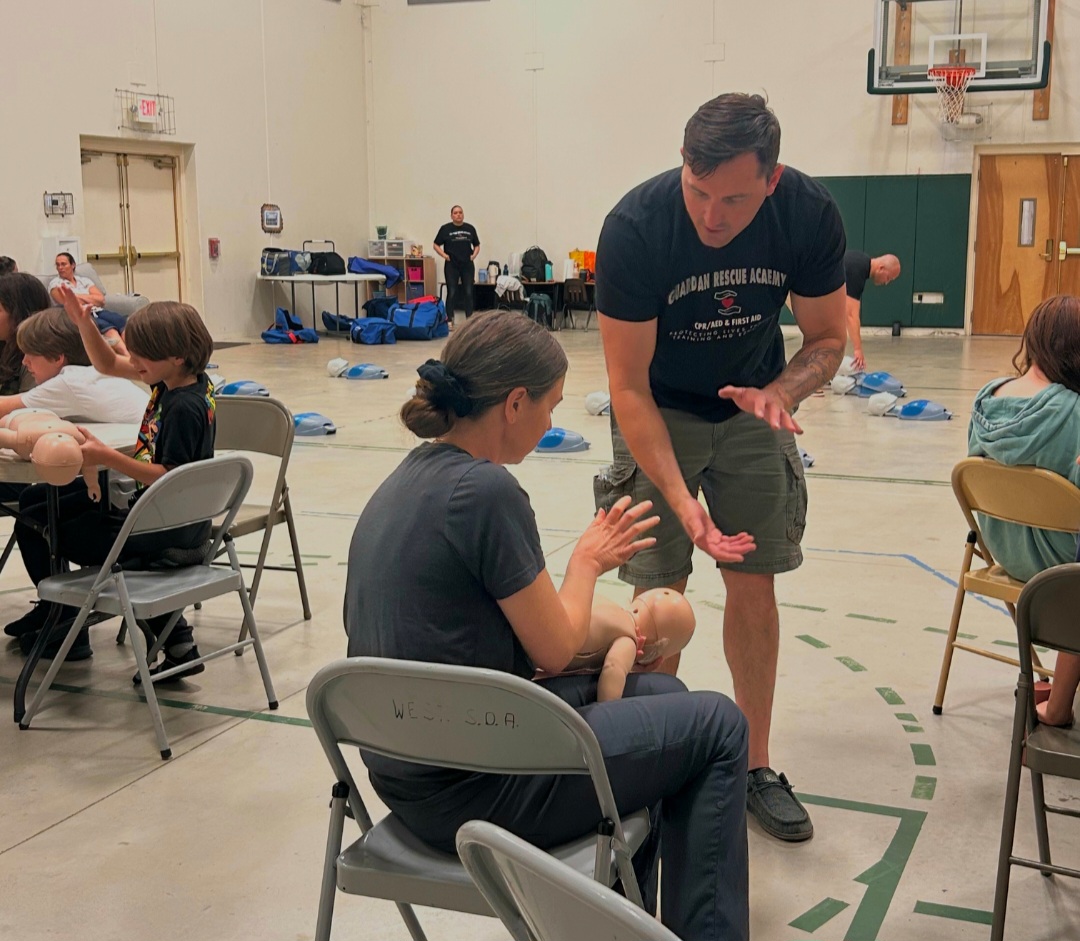 A man interacts with a seated woman in a gymnasium, with children and chairs around. Equipment is visible in the background.