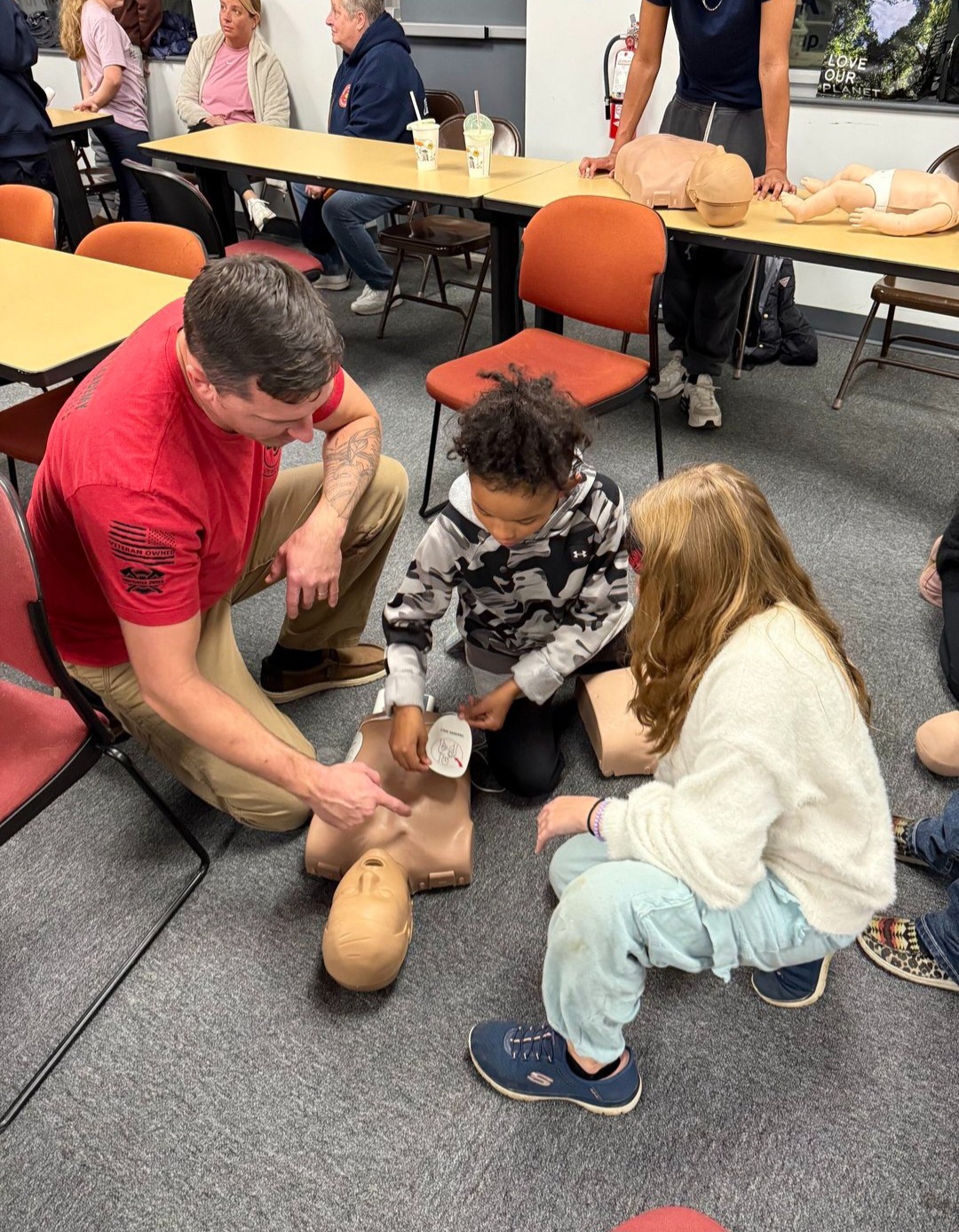 Man demonstrating CPR on a mannequin with two children watching closely in a classroom setting.