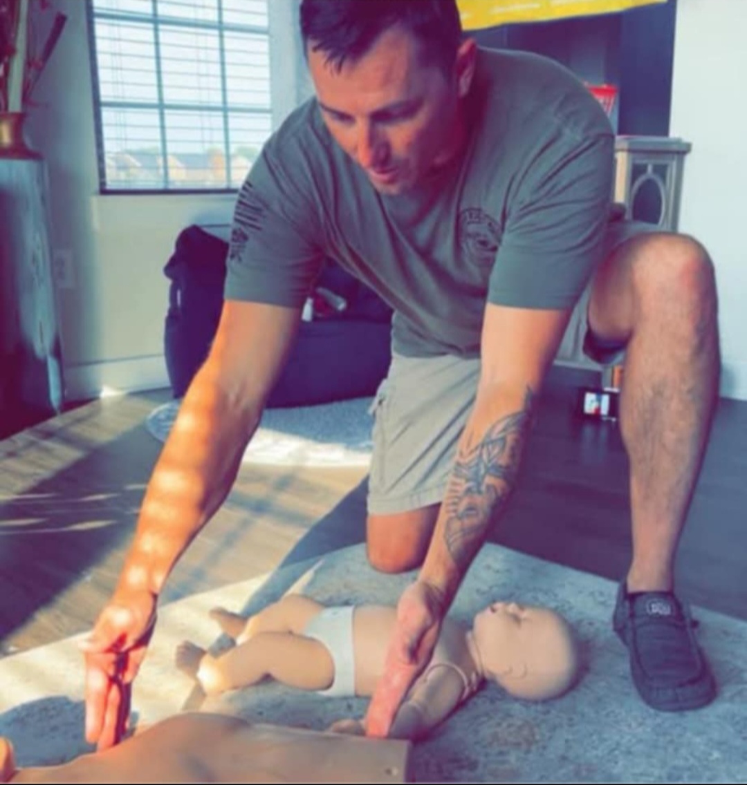 Man practicing CPR on a doll indoors, kneeling on the floor with one hand on the doll's chest and the other on its head.