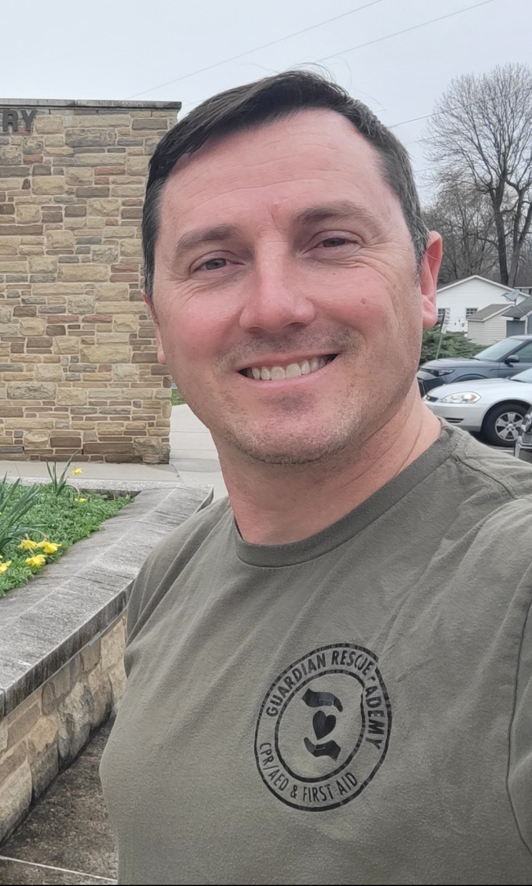 Smiling man outdoors in front of a brick building and parked cars, wearing a gray T-shirt with a circular logo.