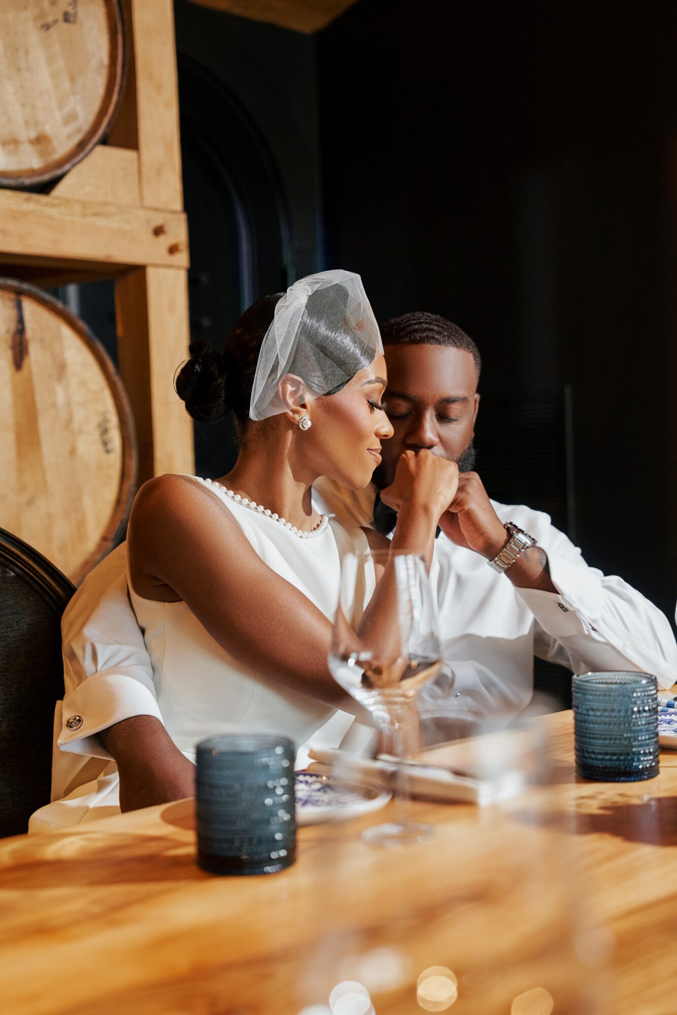 Two people sit at a table with wine glasses and candles, in a setting with wooden barrels in the background.
