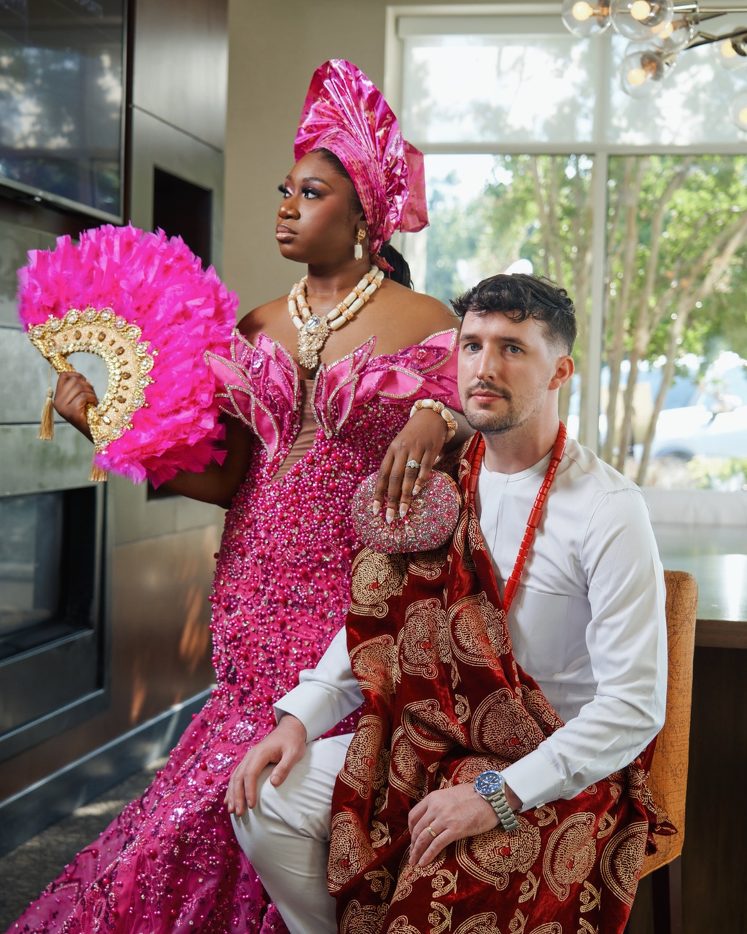 A woman in traditional pink attire with a headwrap and fan, and a man in white with a patterned shawl, indoors.