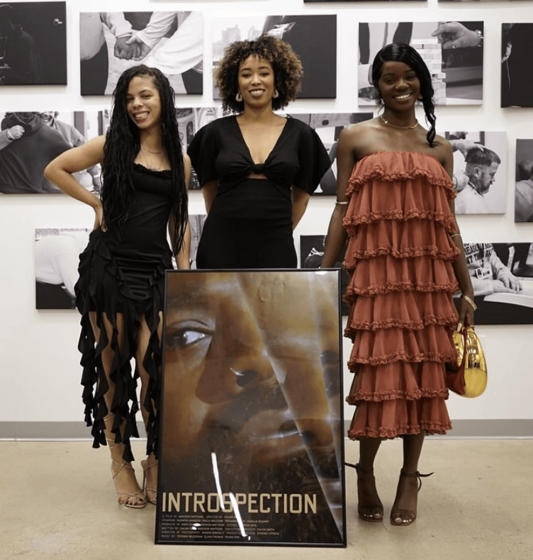 Three women stand in front of a wall with black and white photos, smiling, with a large poster in front of them.