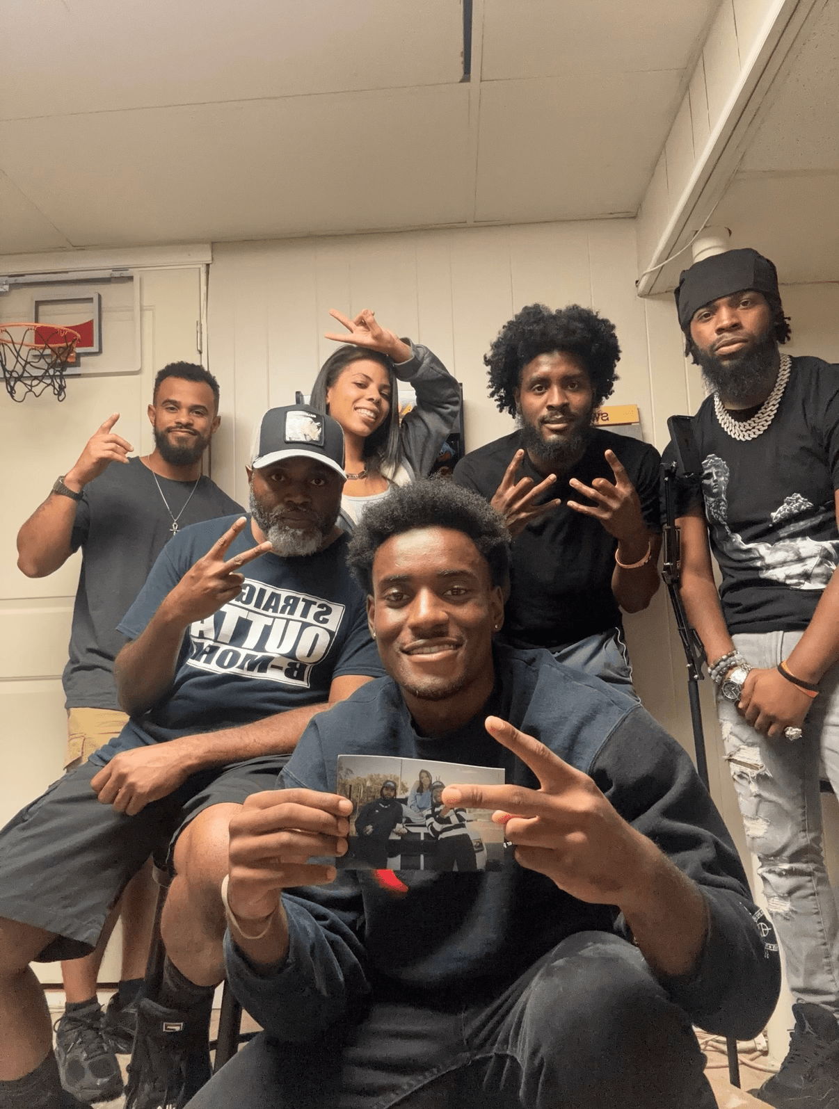 Group of six young people posing indoors, some making peace signs, one holding a phone, smiling at the camera.