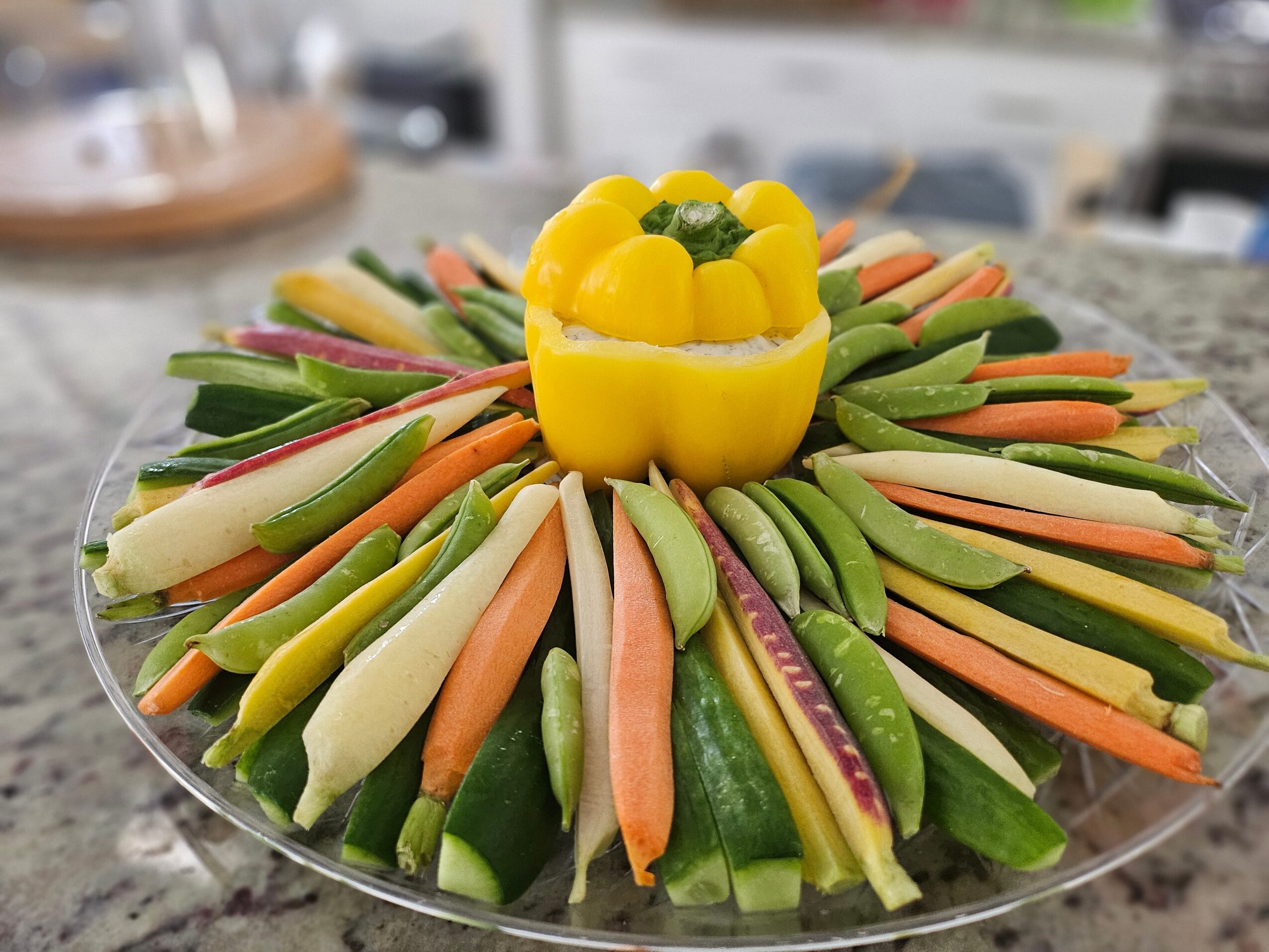Colorful vegetable platter with a yellow bell pepper centerpiece, surrounded by sliced cucumbers, carrots, and other vegetables, on a clear plate.