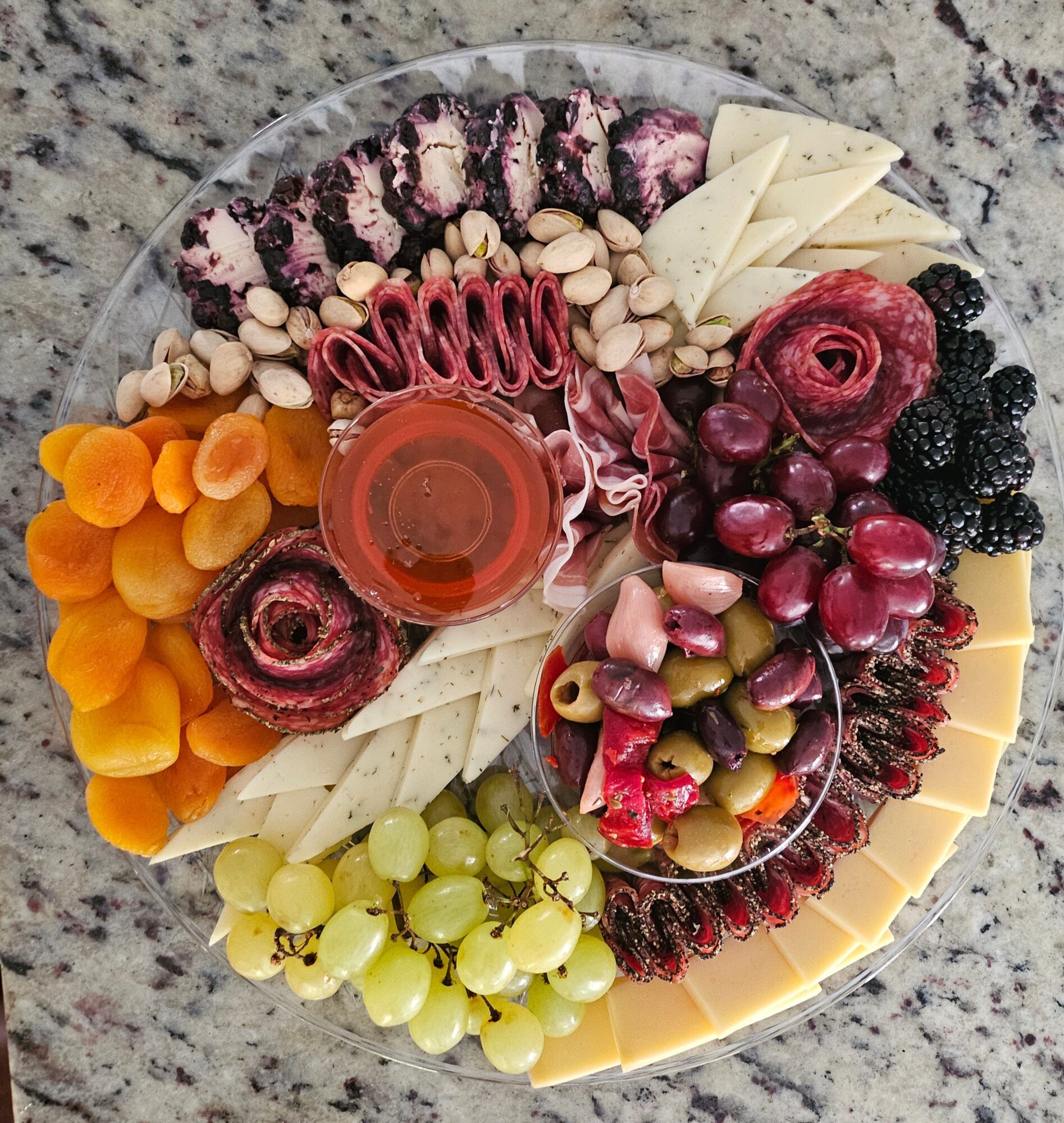 Round platter with assorted cheeses, grapes, dried apricots, nuts, and cured meats on a granite surface.