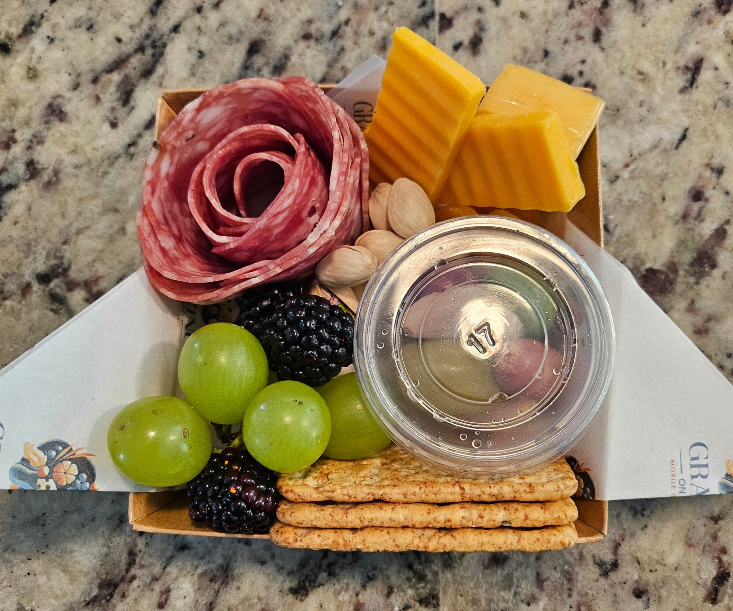 Assorted cheese, sliced meat, green grapes, blackberries, crackers, pistachios, and a small container of dip on a white plate.