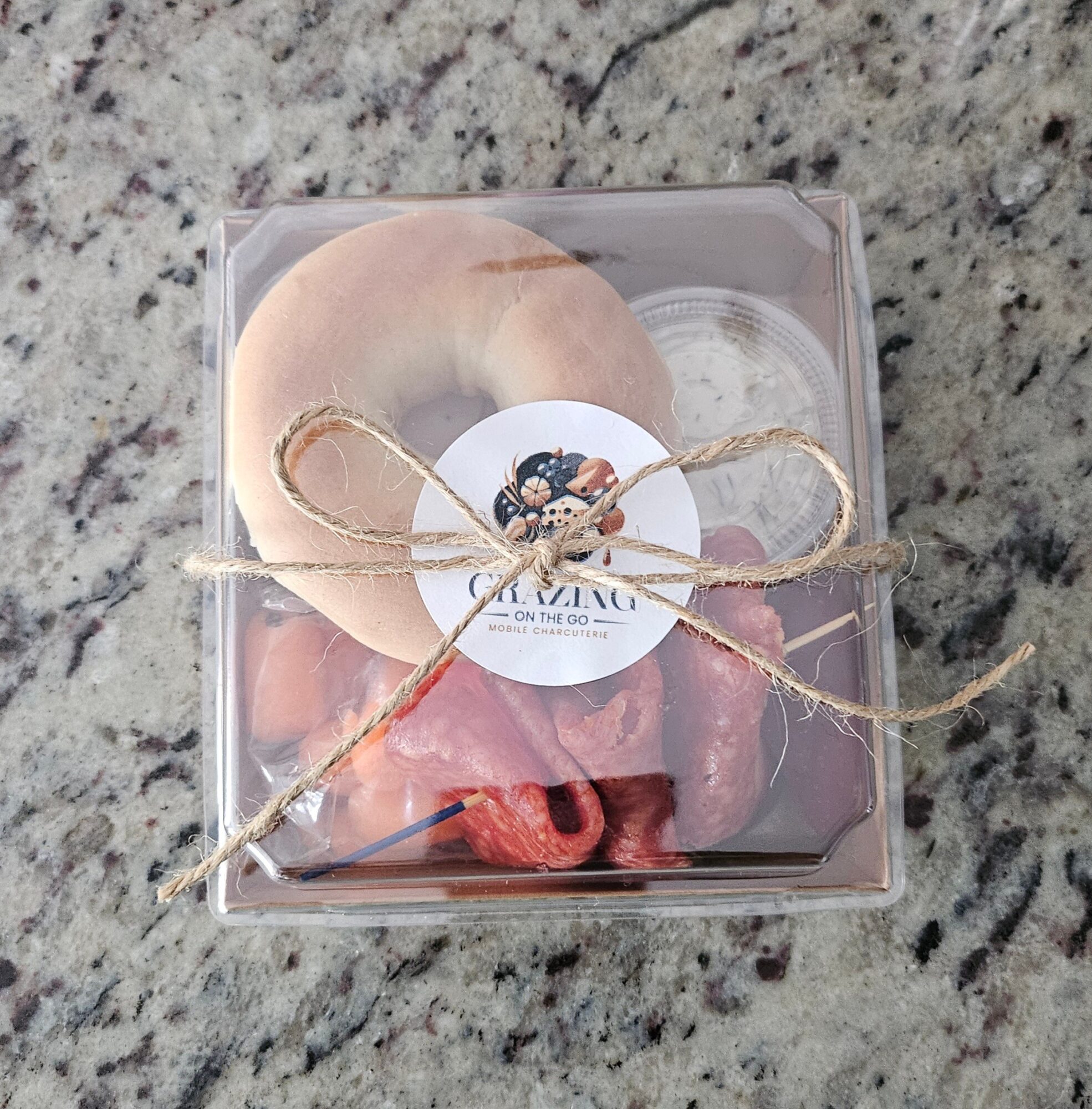 Clear box with a round cookie, a pink item, and a small container, tied with a gold string, on a granite surface.