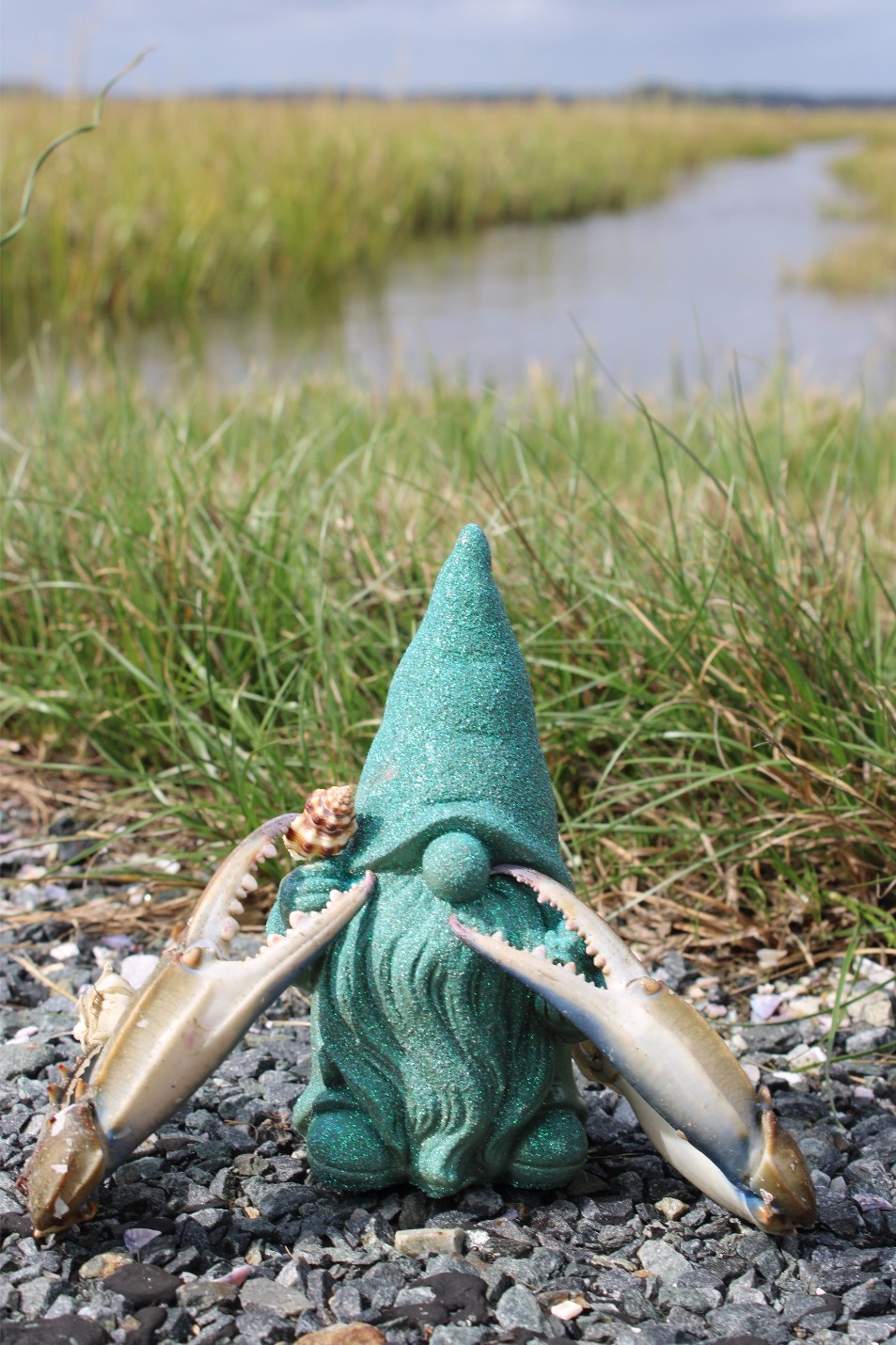 A garden gnome with a tall, pointed hat, surrounded by two white fish, outdoors near a body of water and grassy landscape.