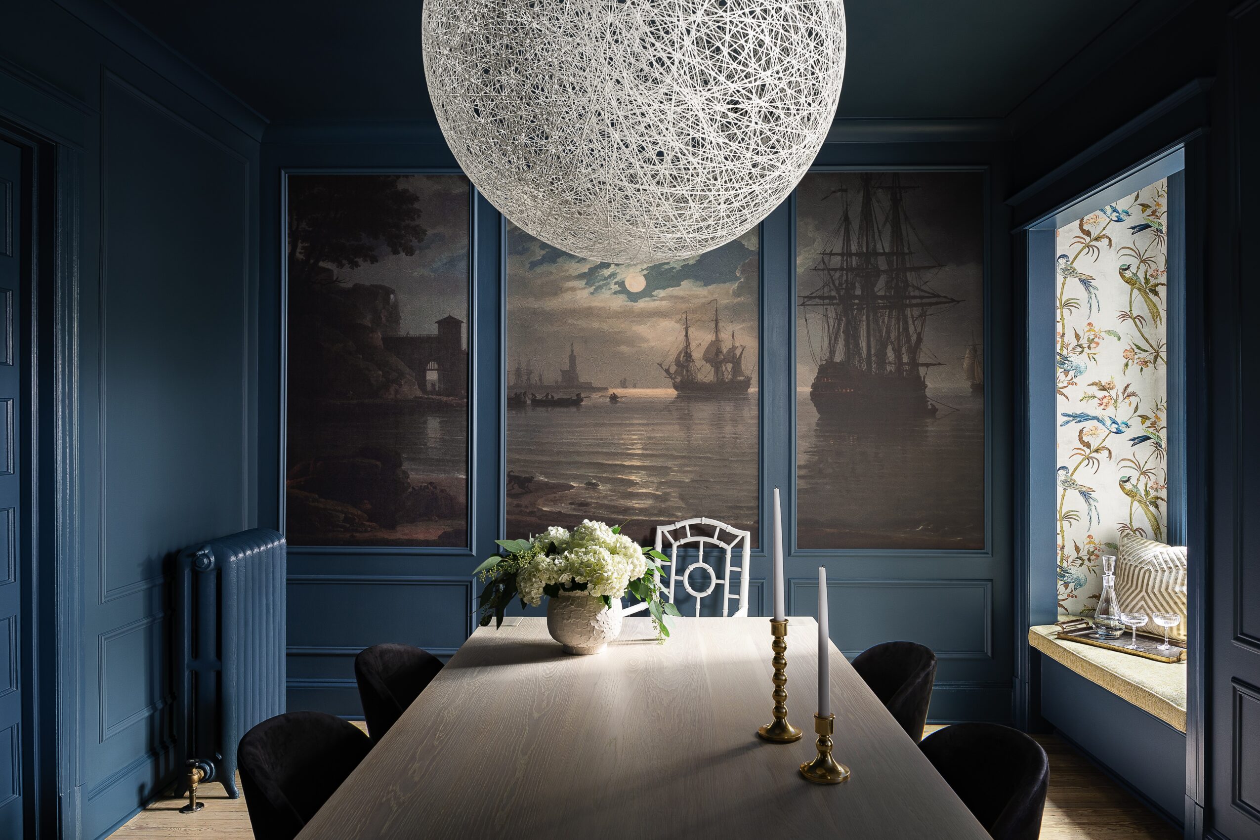 Room with dark blue walls, large window, and a round textured ceiling light fixture, with a table and decorative items.